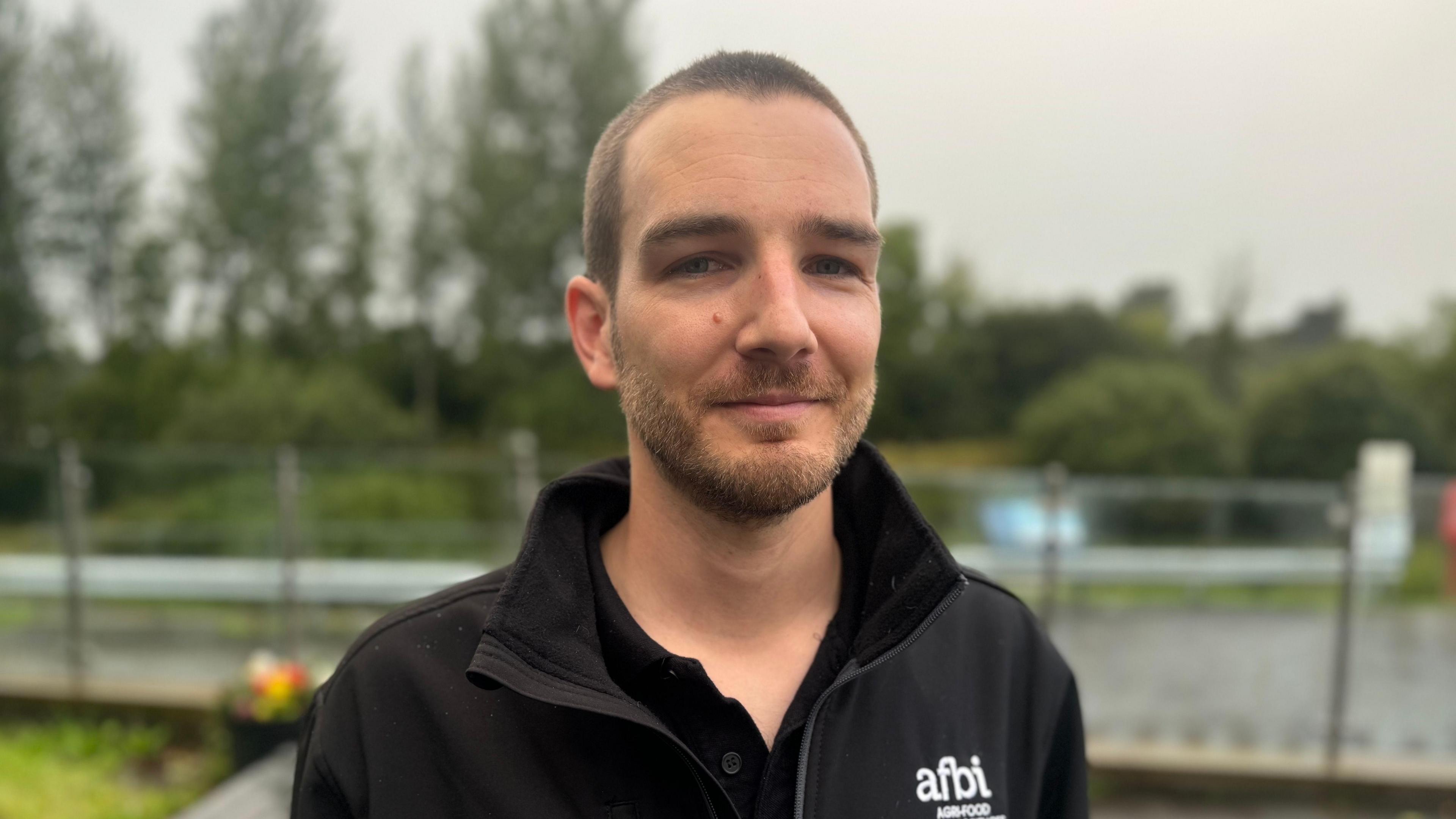 A man is smiling directly into the camera. He is wearing a black fleece with branding from the Agri-Food and Biosciences Institute and a black polo shirt. He has dark-coloured hair cut short and a brown beard. Our of focus in the background you can see trees and a fence. 