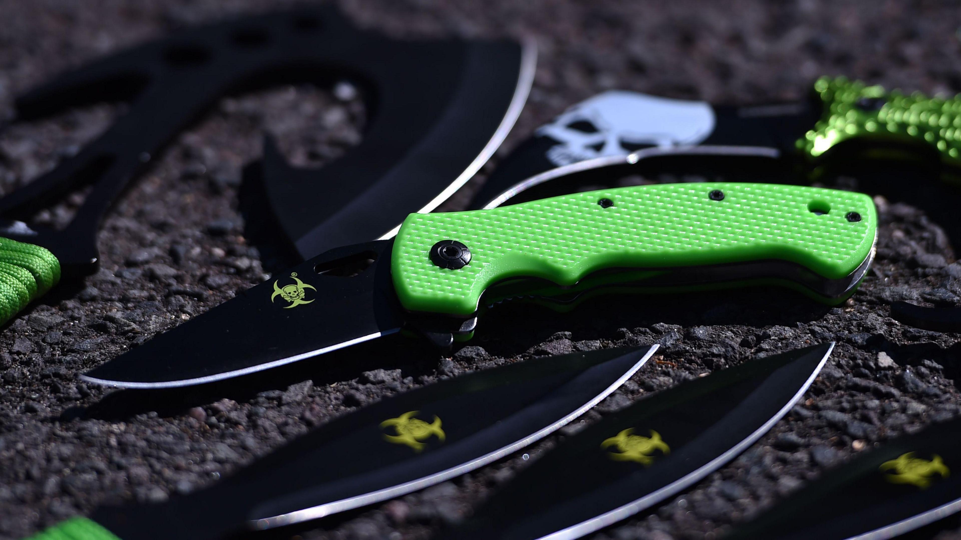 A zombie-style knife with a green handle and a skull an cross bone in green on the blade sits on the ground, with other, similar blades surrounding it. 