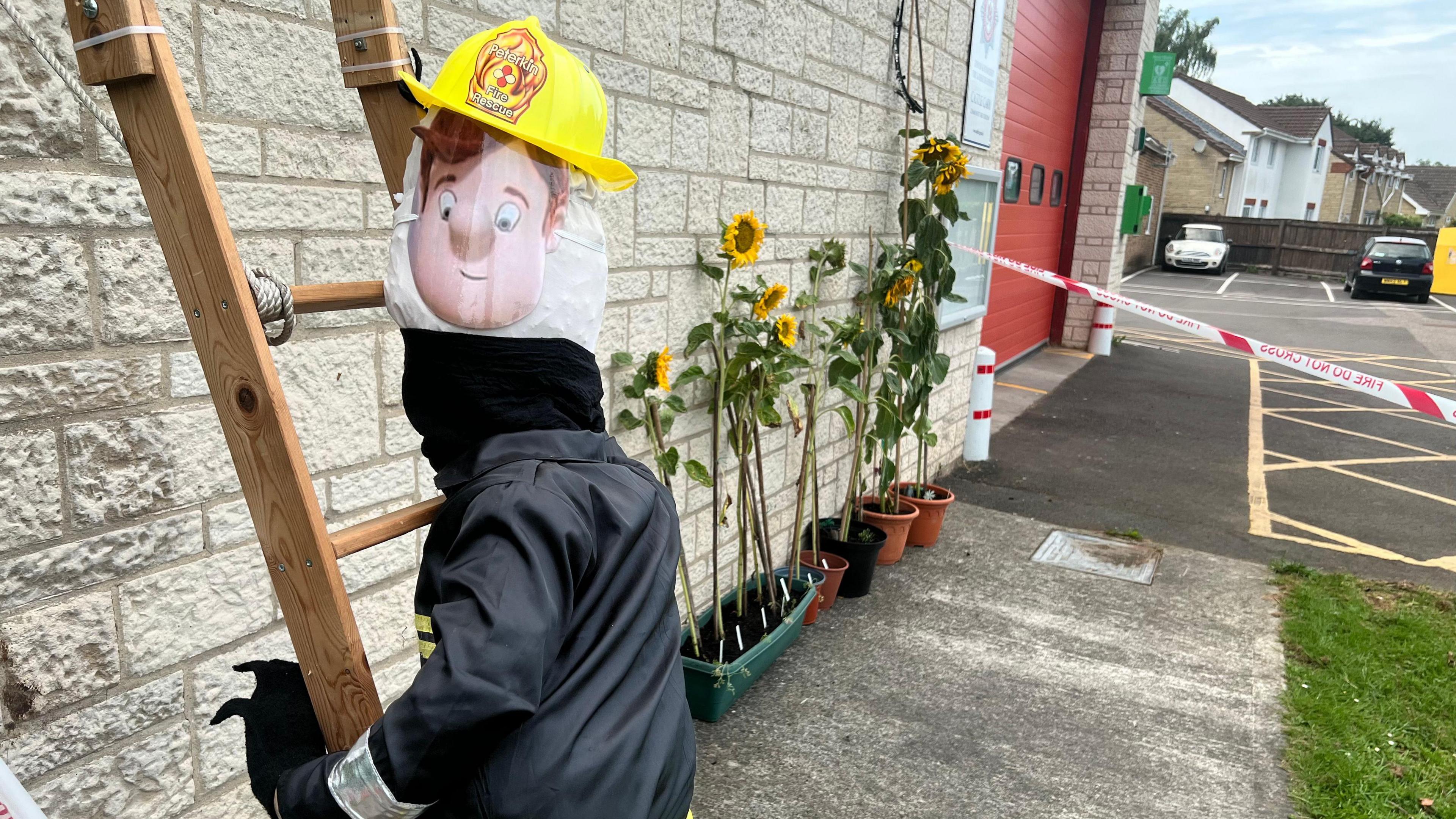 A Fireman Sam scarecrow climbing a ladder next to some sunflowers