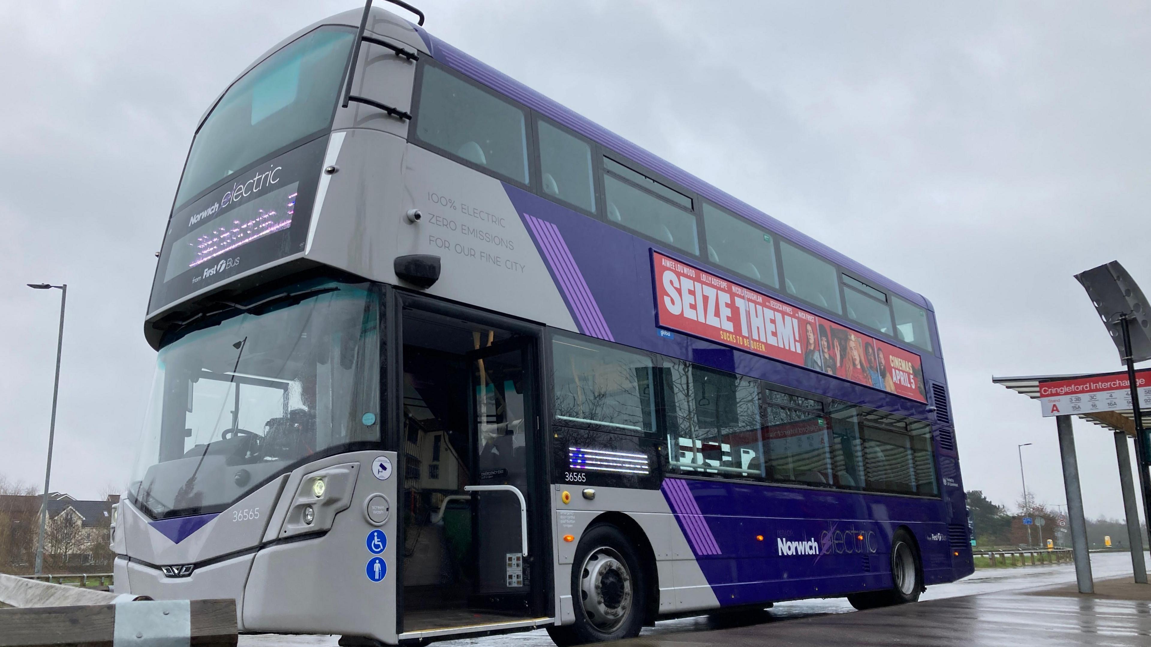 A purple and grey electric double-decker bus parked at a bus stop on a rainy day. The bus door is open and a promotional sign on the side of the bus reads 'seize them'.