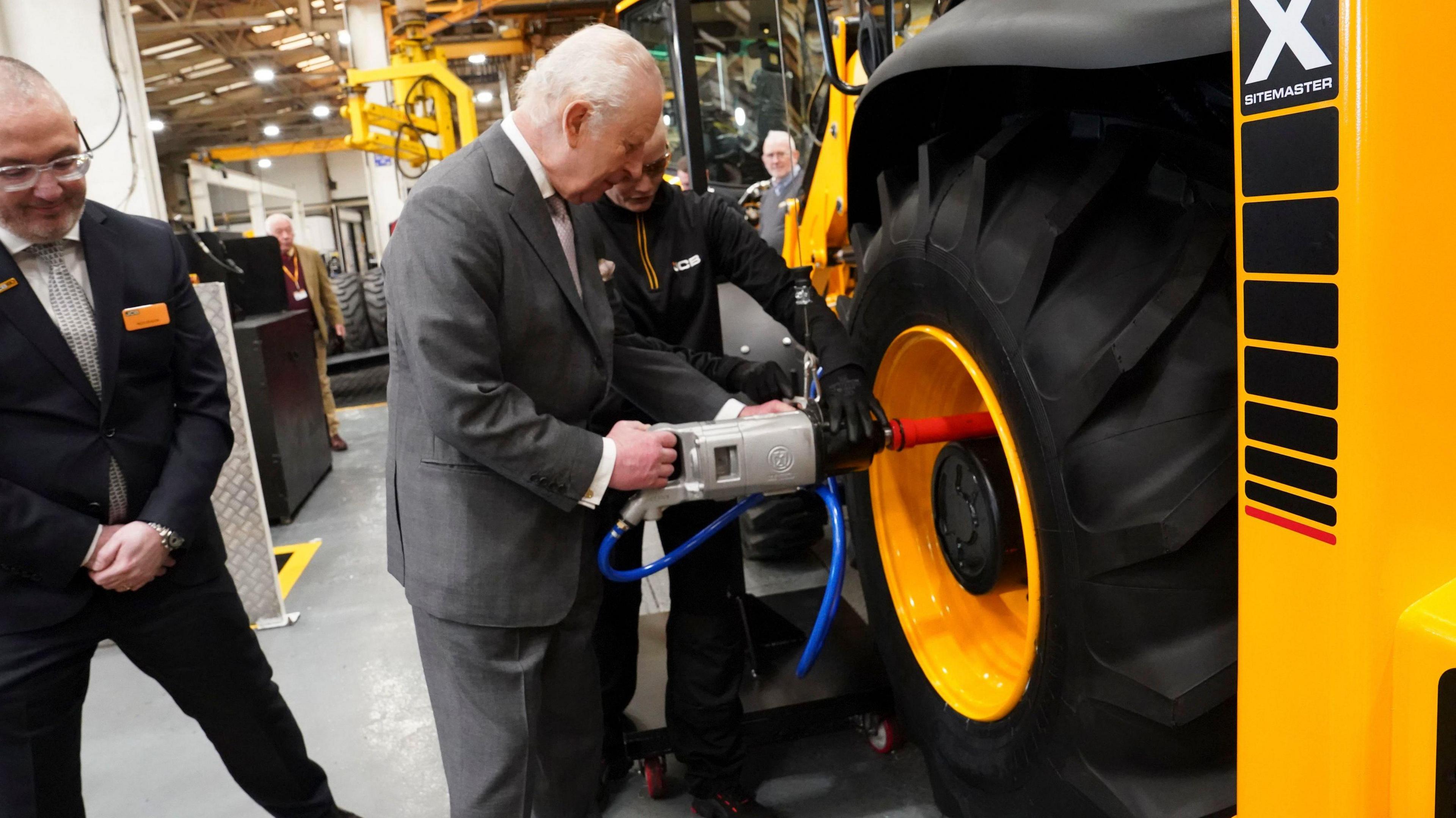 King Charles wearing a grey suit stands next to two other men and next to a large tyre fitted to a large yellow vehicle. He is holding a piece of equipment as he fits the tyre to the vehicle.