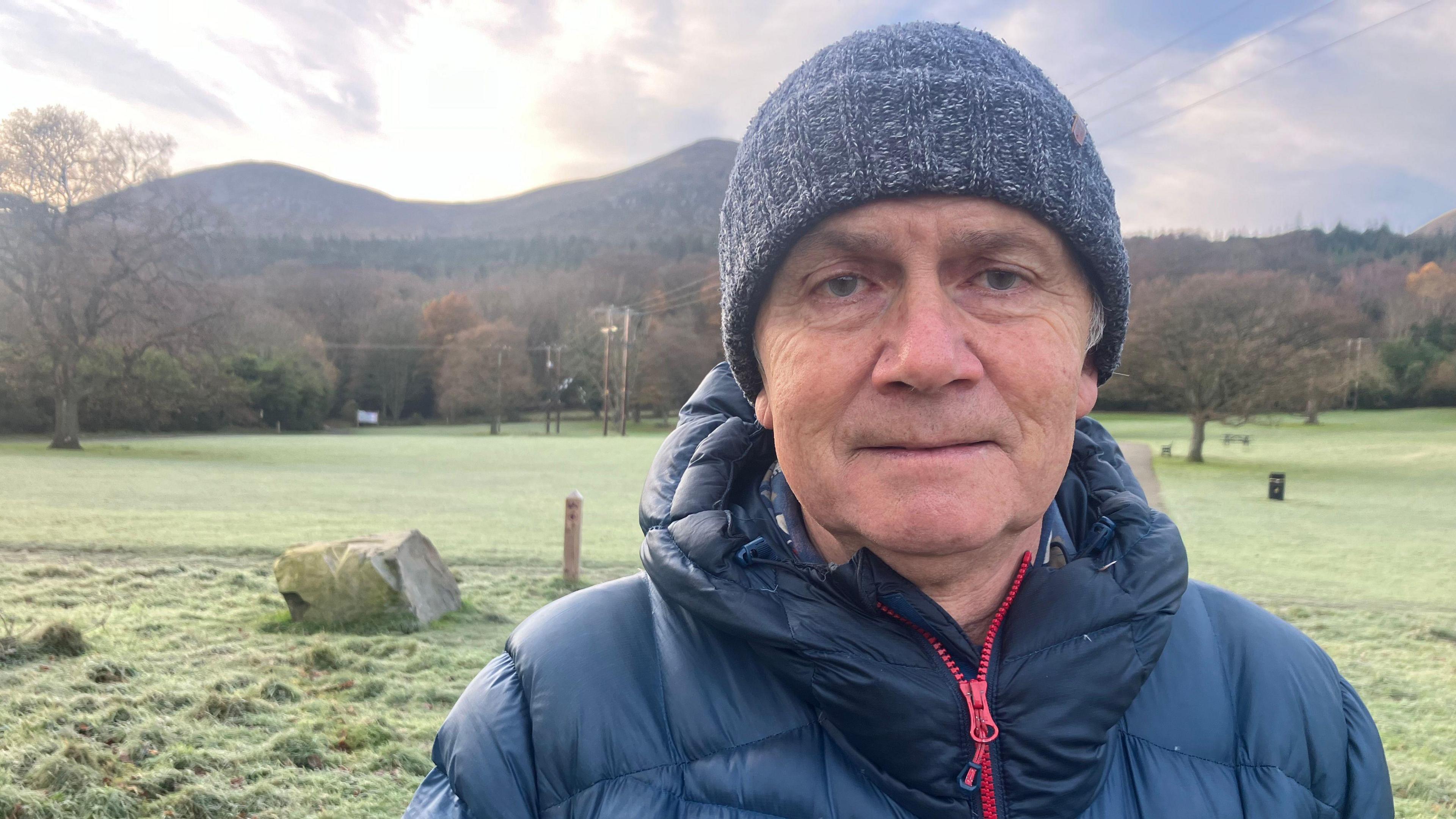 A man wearing a blue zip up coat is looking toward the camera. He is wearing a blue knit hat. In the distance is a large mountain range. 