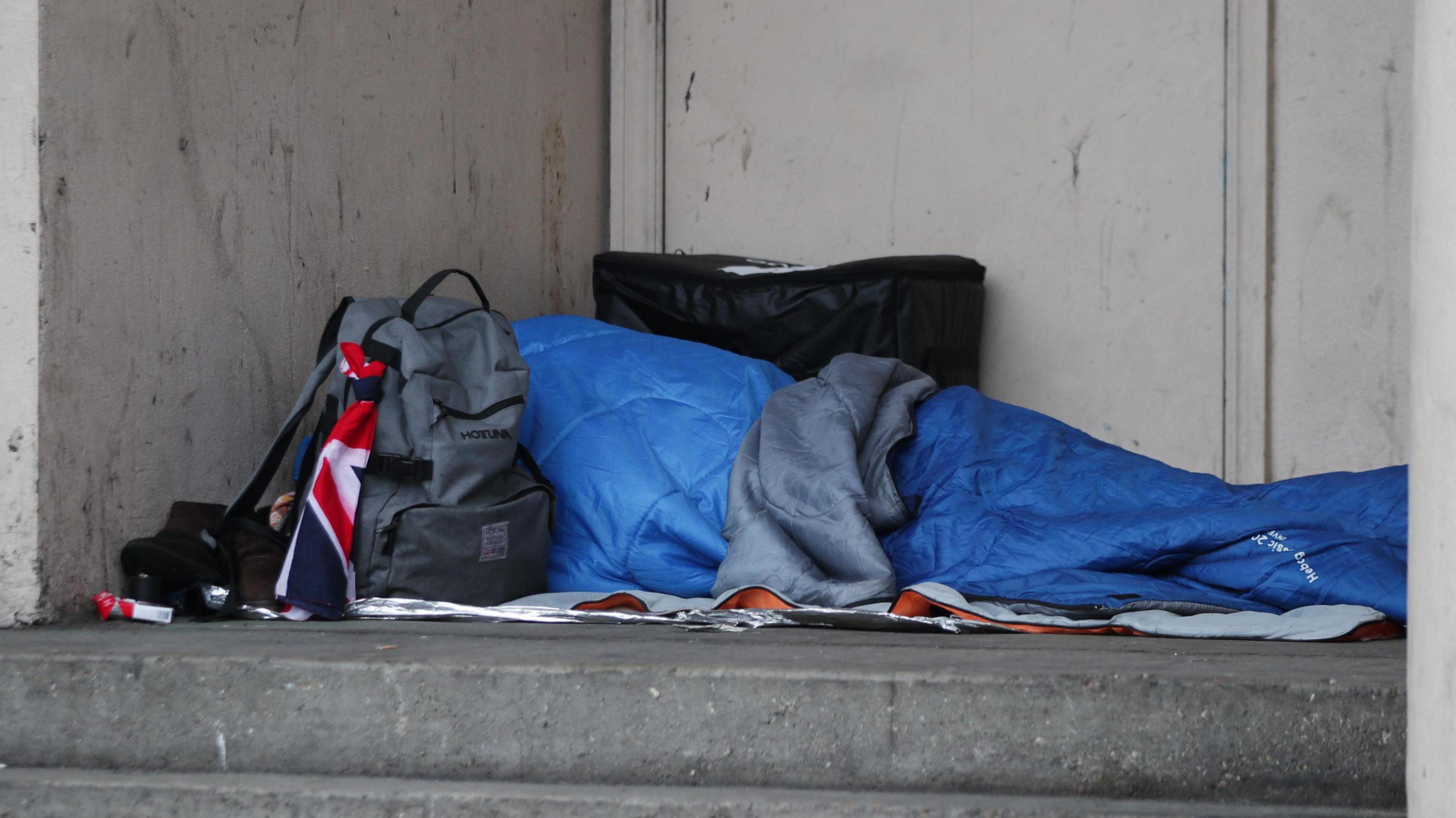 Person, who is not visible, asleep in a blue sleeping bag on a grey step in front of a grey wall.