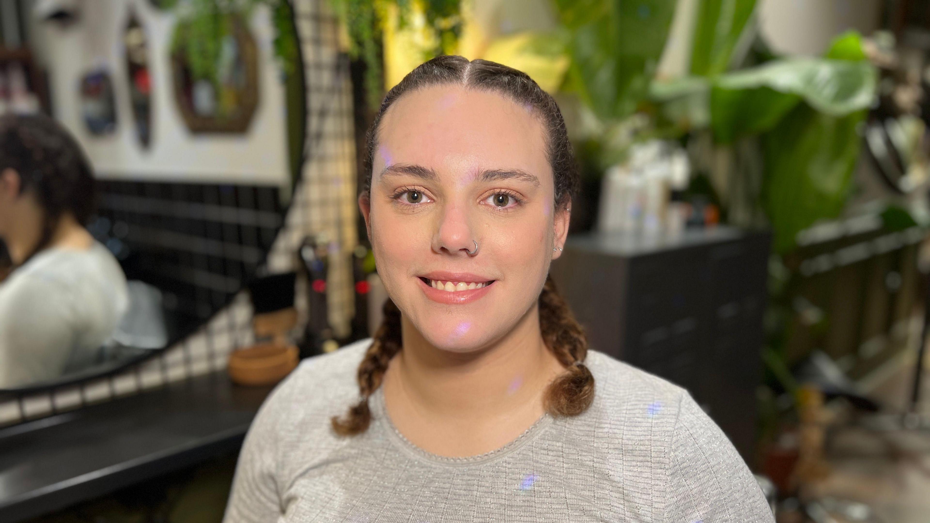 Annabelle Sonmez, a 26-year-old woman with a nose piercing. She is sitting in a hair salon with plaits in her curly hair. She is wearing a grey long-sleeved T-shirt and is smiling.