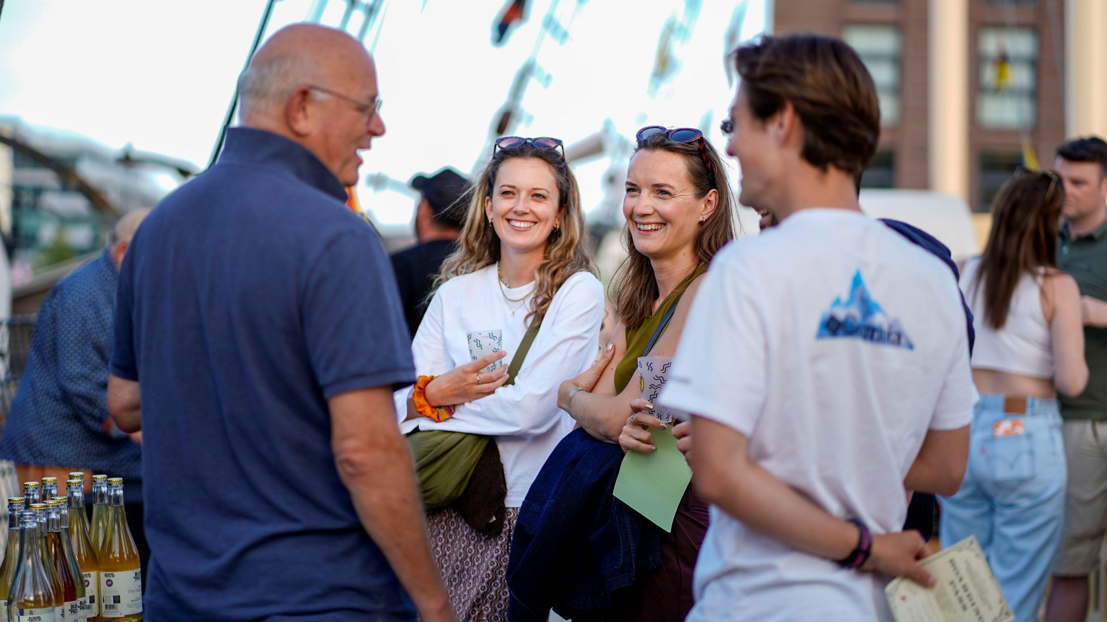 A group of four people, two men and two women, smile as they talk at a cider event on the SS Great Britain in Bristol. The rigging of the ship is visible, but not in focus, in the background