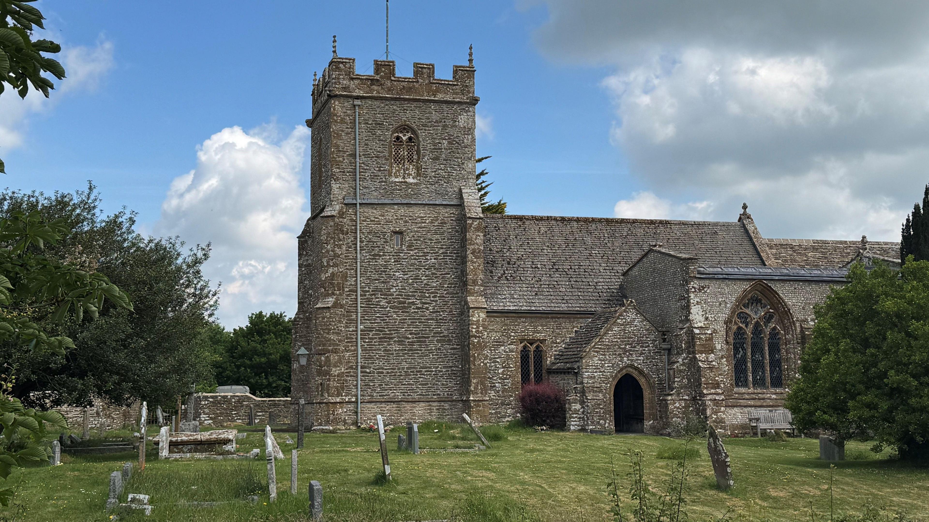 Exterior of St Mary's church. A medieval stone church with a crenelated tower but no spire. The building sits within the grass churchyard where there are a number of upright and wonky gravestones.