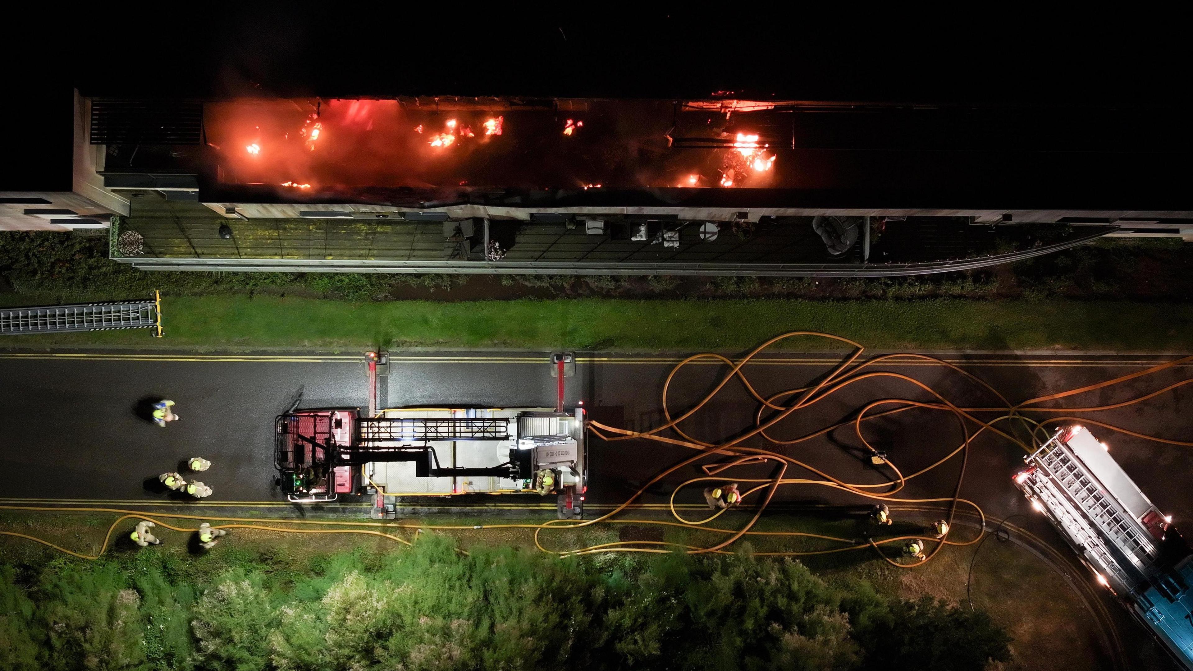 A bird's-eye view of a fire engine and firefighters at the base of the building, with flames visible on the roof. 