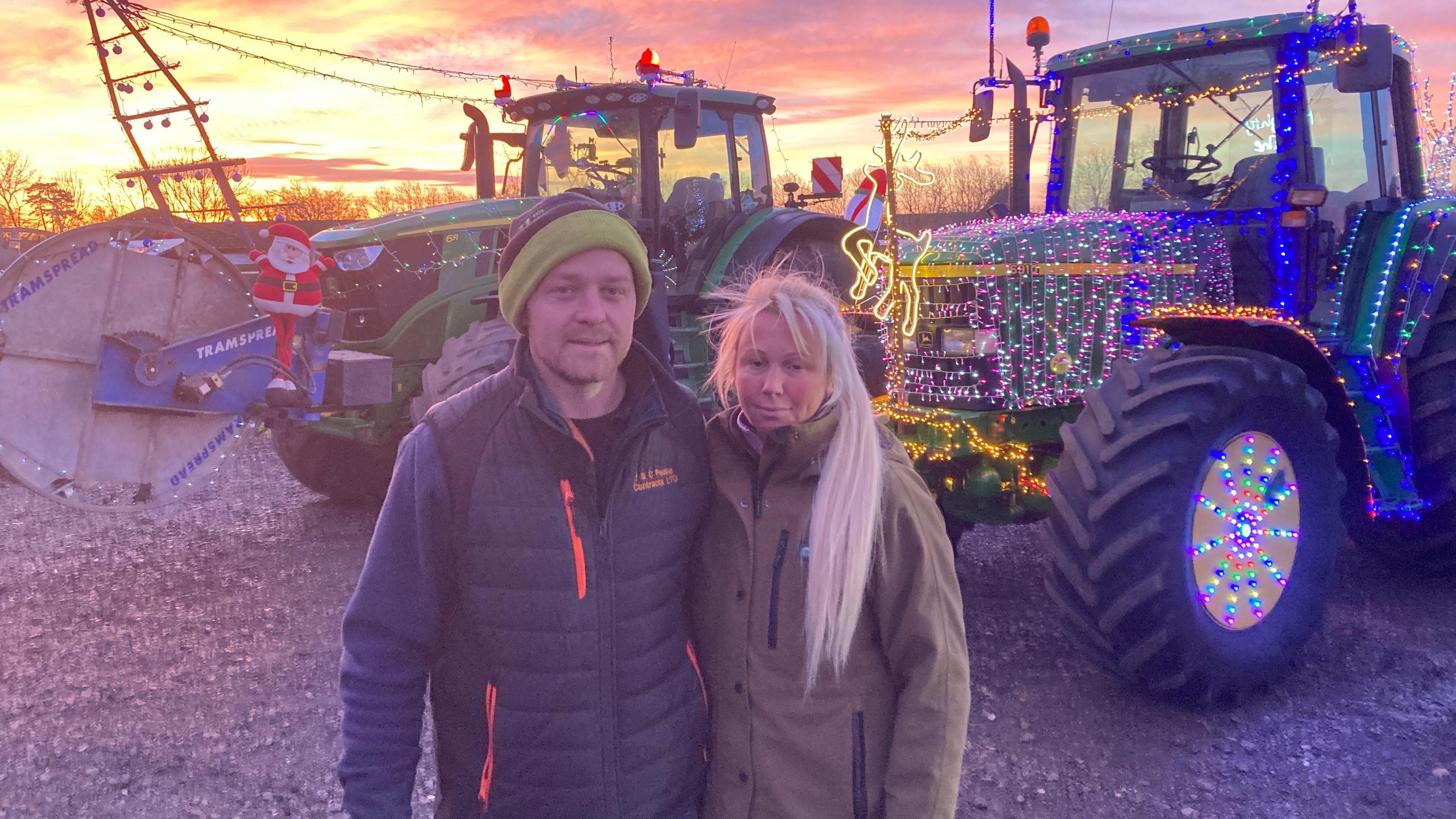 A man and a woman stand next to each other in a field. Behind them are two tractors - one blue and one green - both decorate din Christmas lights. The sunrise is also visible on the horizon.