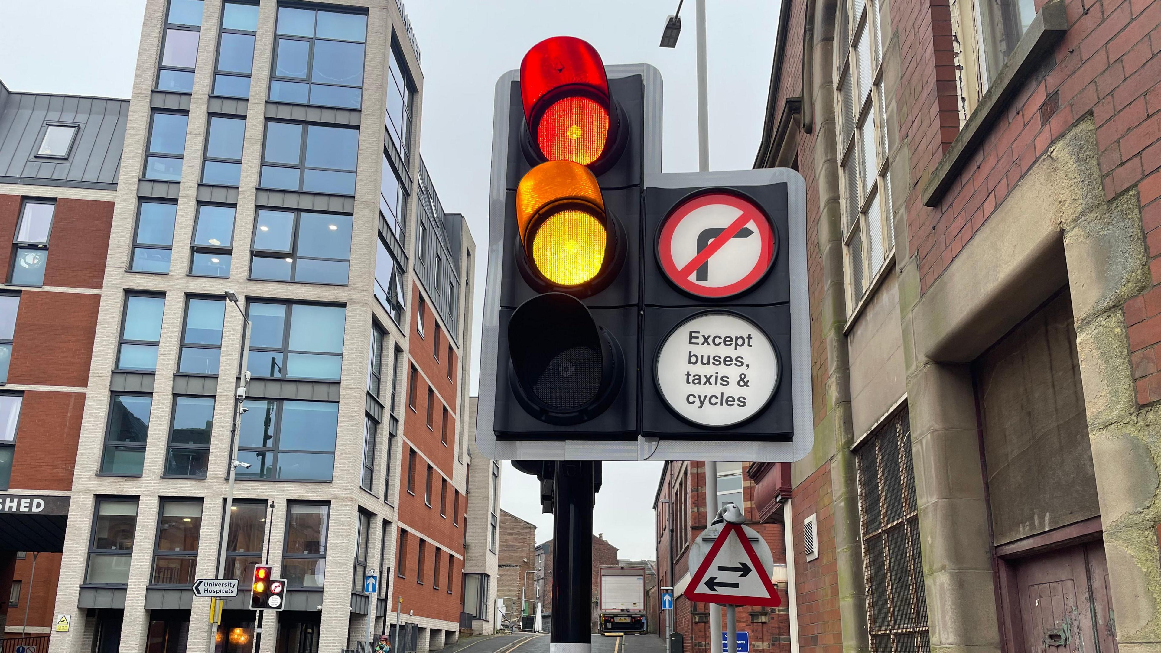 A traffic light about to turn from amber to green is in the foreground, it has a traffic sign indicating no right turn except for buses, taxis and cycles.  In the background there is another sign that indicates traffic travelling in both directions. Also in the background is a student halls called the Tramshed and some other red-brick buildings.