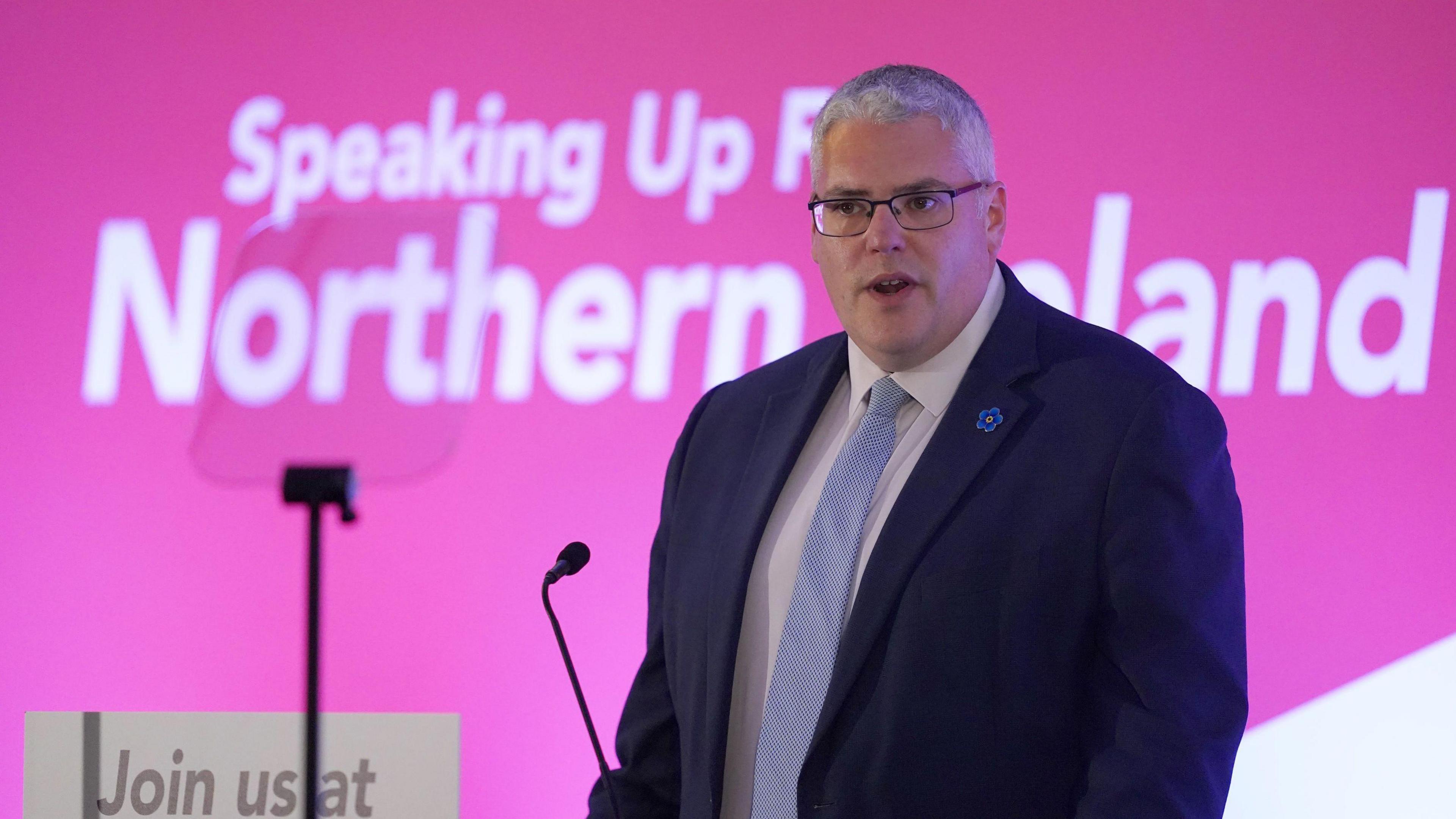 Gavin Robinson speaking at a microphone with a teleprompter in front of him. He is wearing a suit and tie. Behind him in large lettering it says "Speaking up for Northern Ireland"