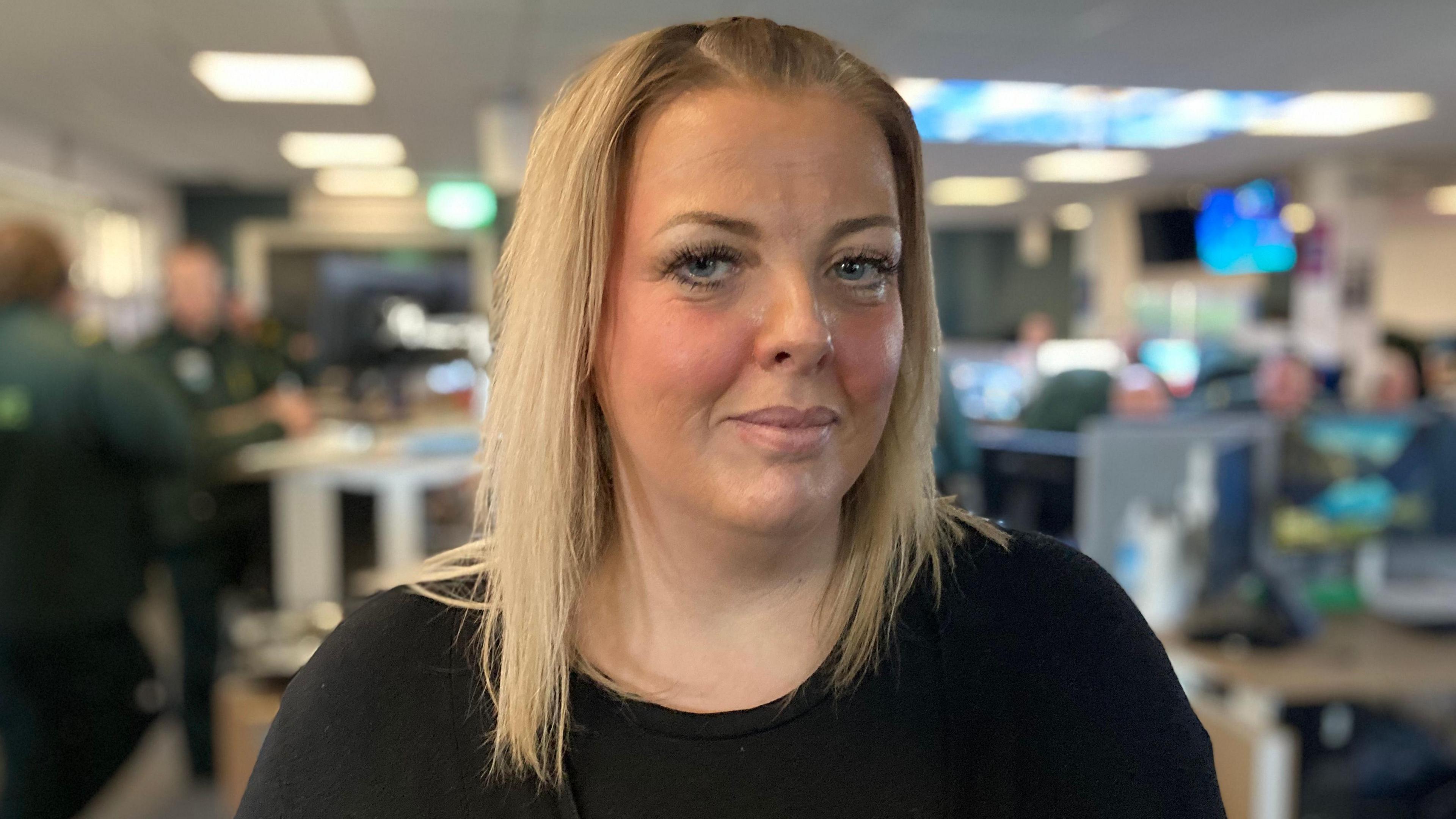 Anne-Marie Scott - a woman with shoulder-length blonde hair is smiling at the camera. She is wearing a black, long-sleeved top. She is standing in the middle of a call centre, but the background is blurred.