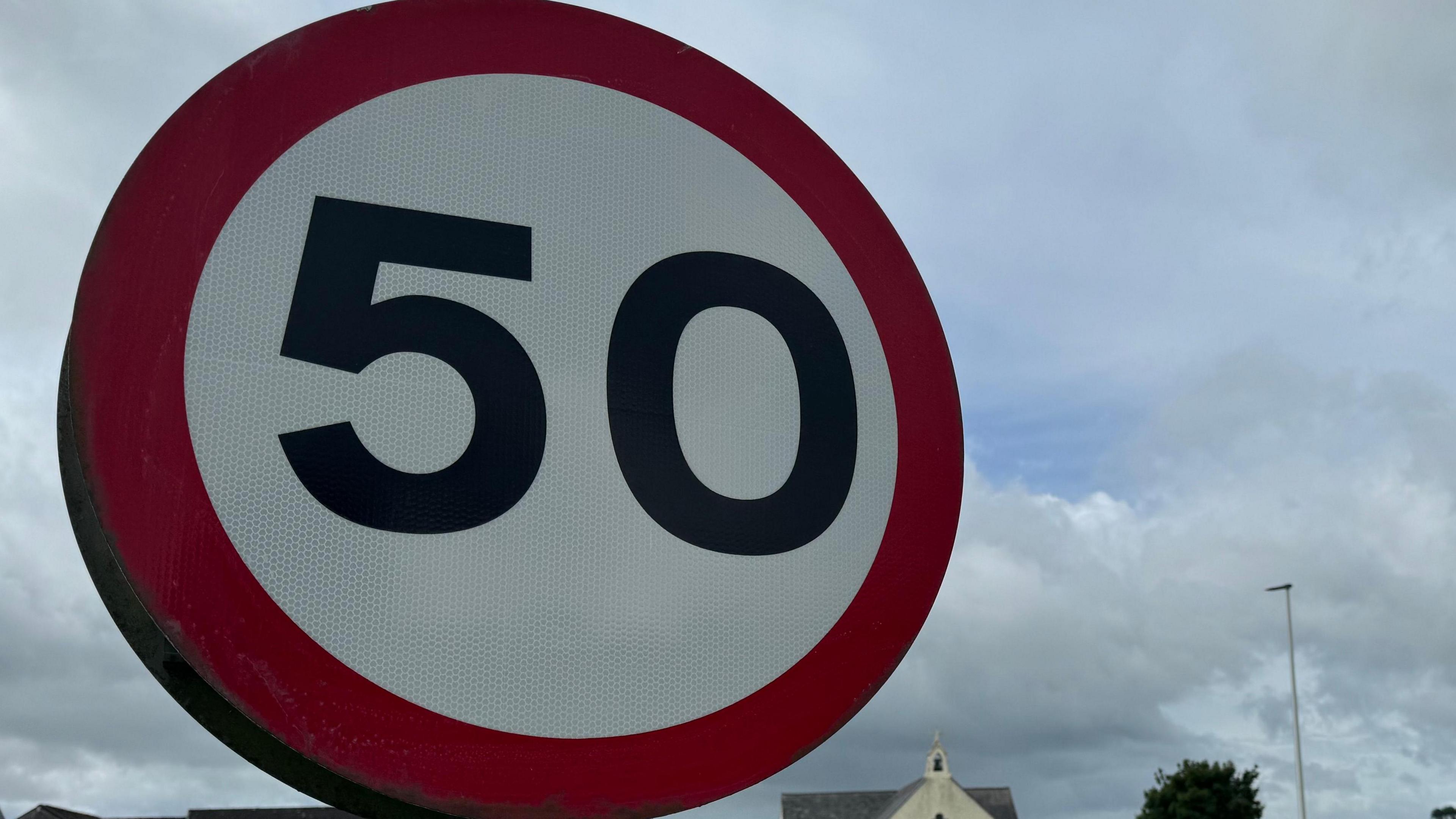 On the left of the image is a circle which has a red boarder around it. The middle part is white, and has a black number 50 written on it. It is a road sign that is on the side of a road. 