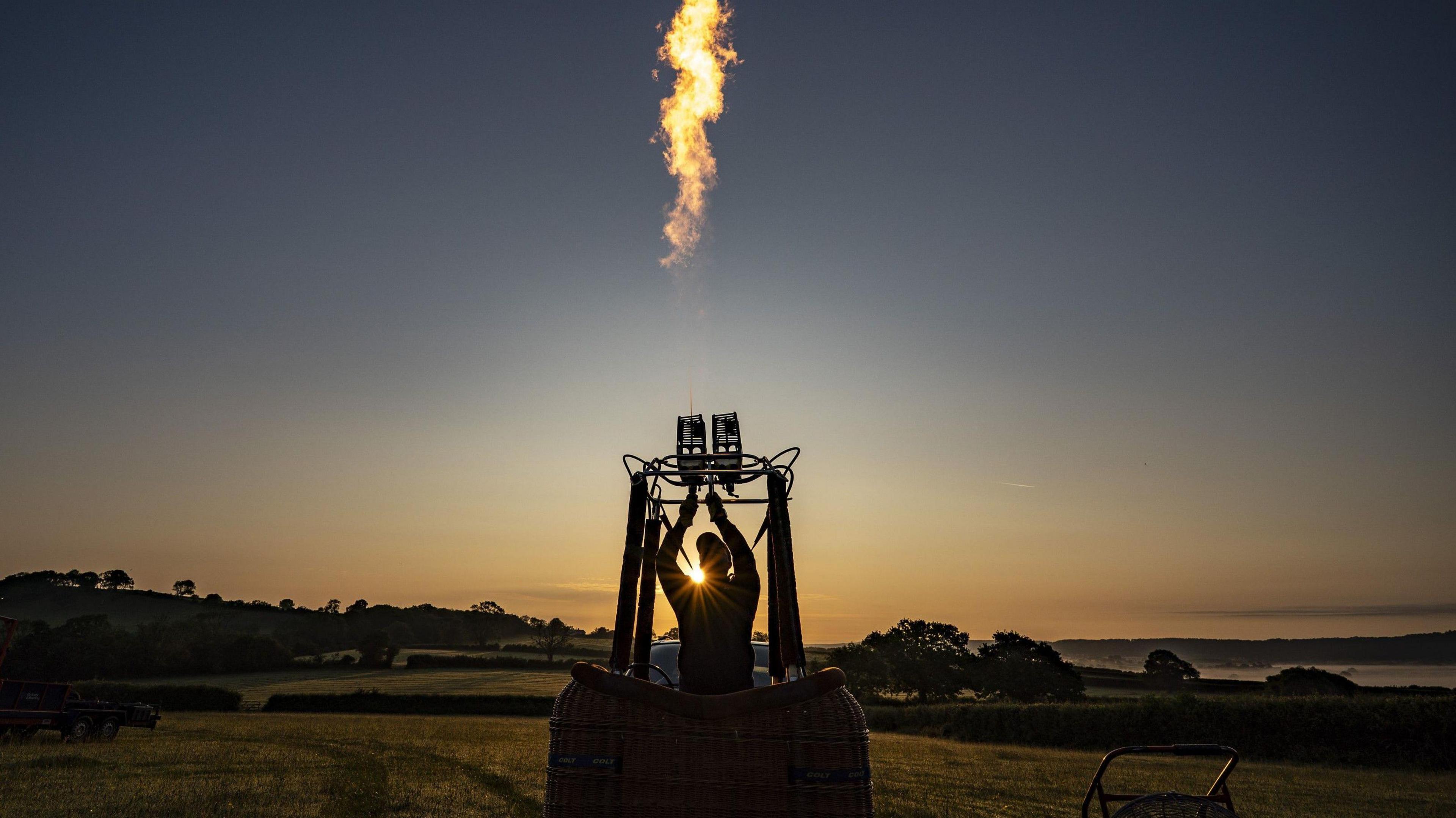 A person standing in a hot air balloon basket fires up the burners against an early morning sky of dark blue and orange