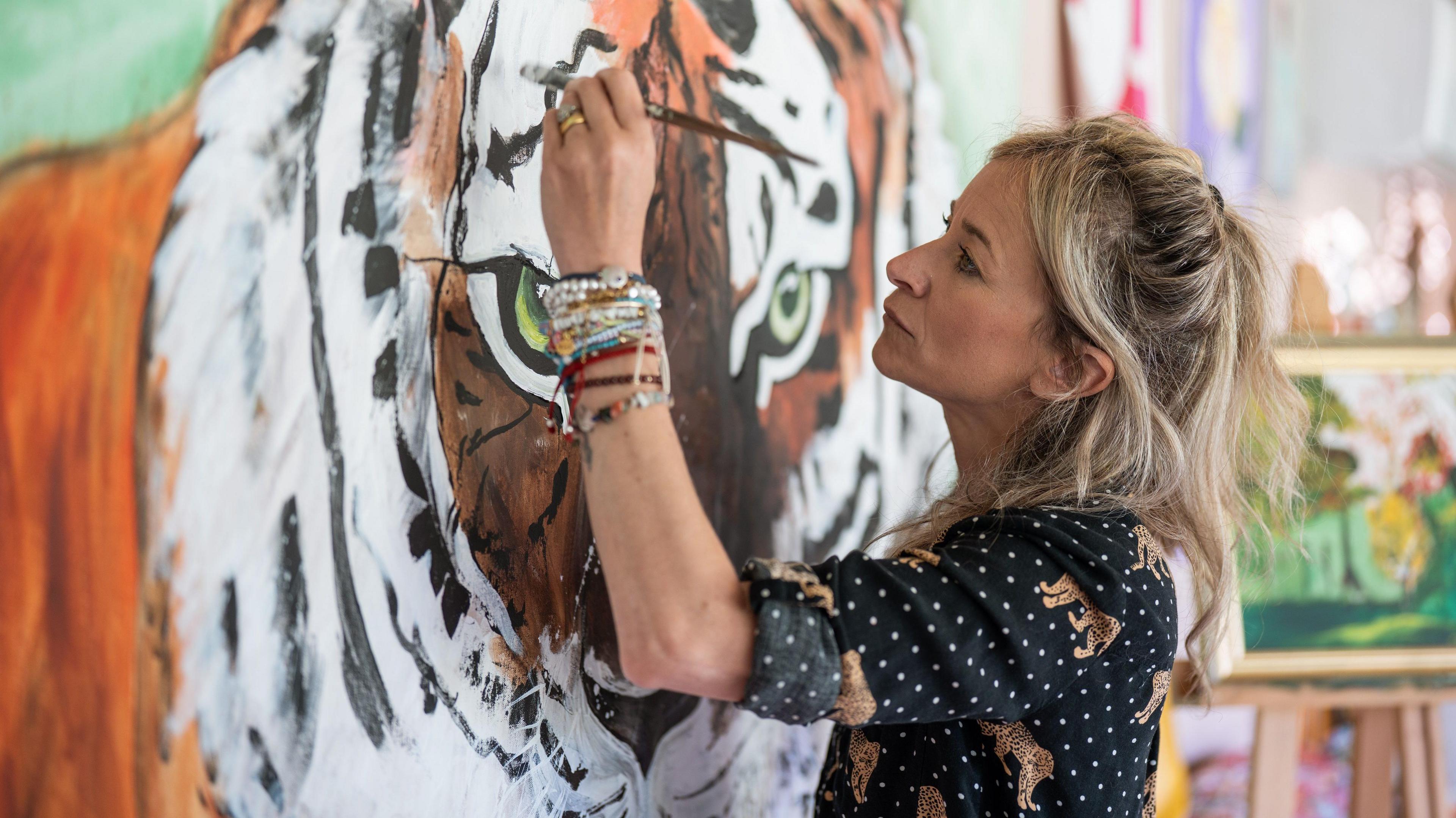 A woman is painting a tiger portrait surrounded by artwork