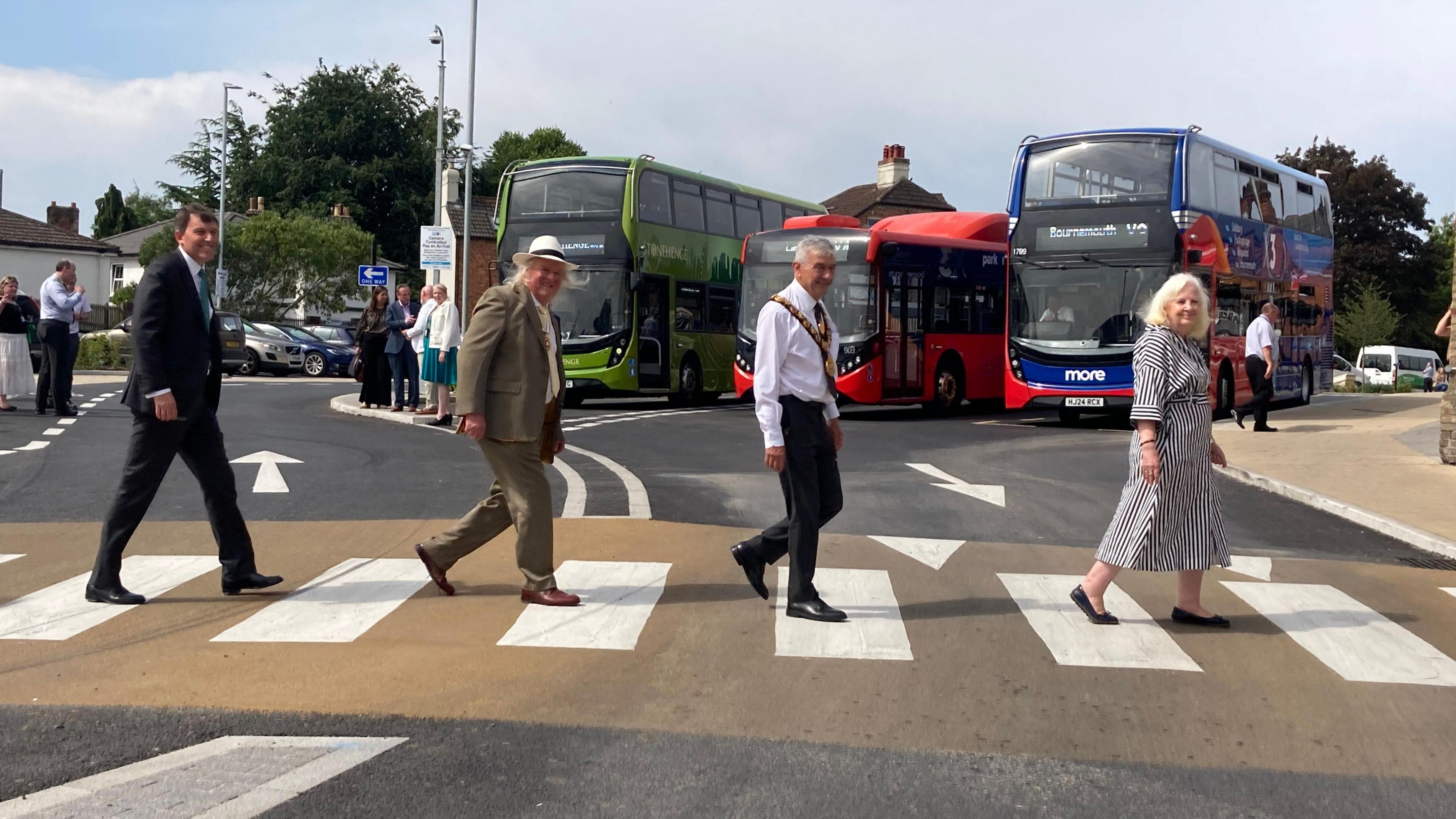 Four people walking on a public crossing, emulating the Beatles album cover. Buses and people in the background.