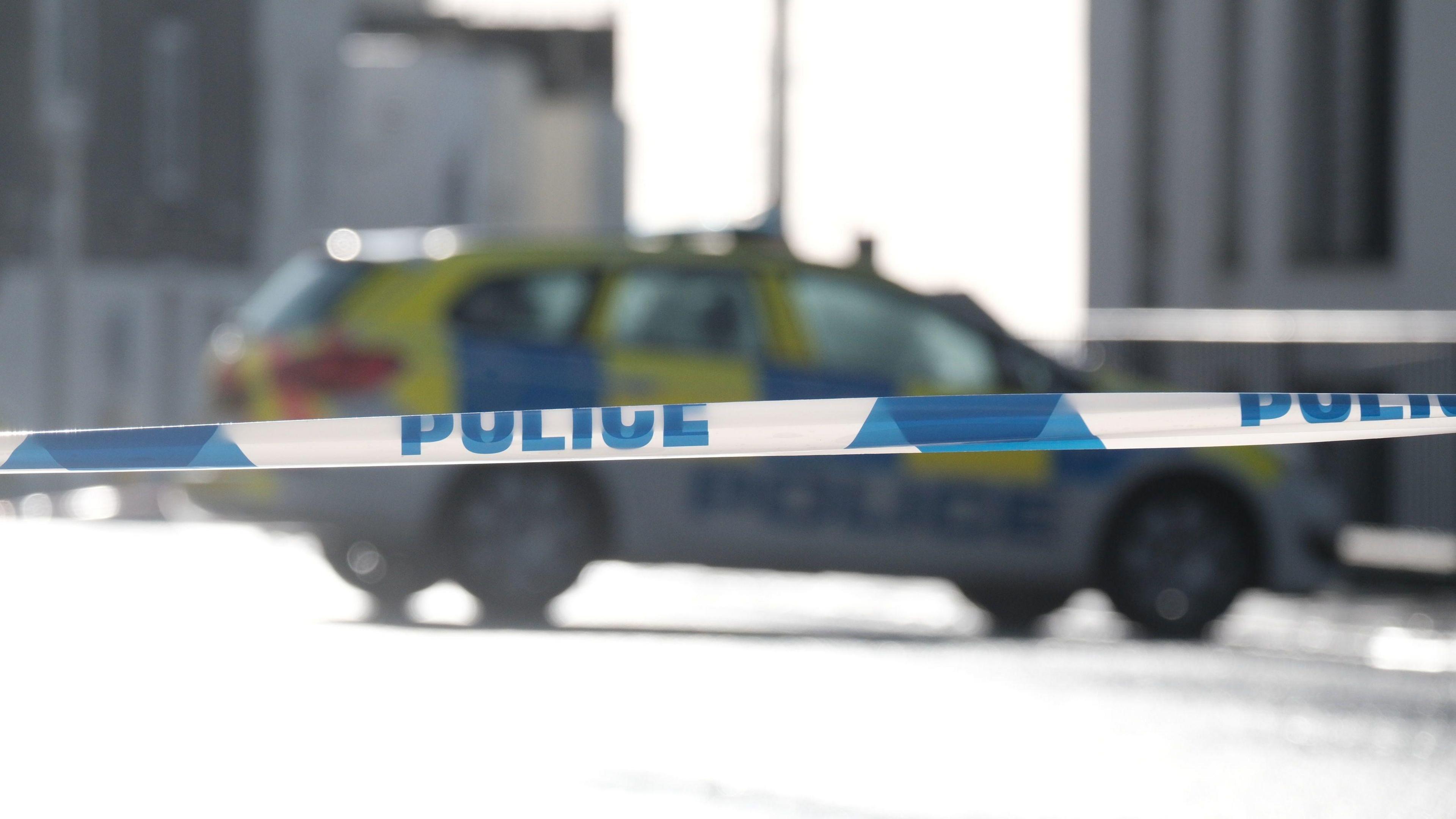 A blurred police car parked on a road in the distance behind police tape.