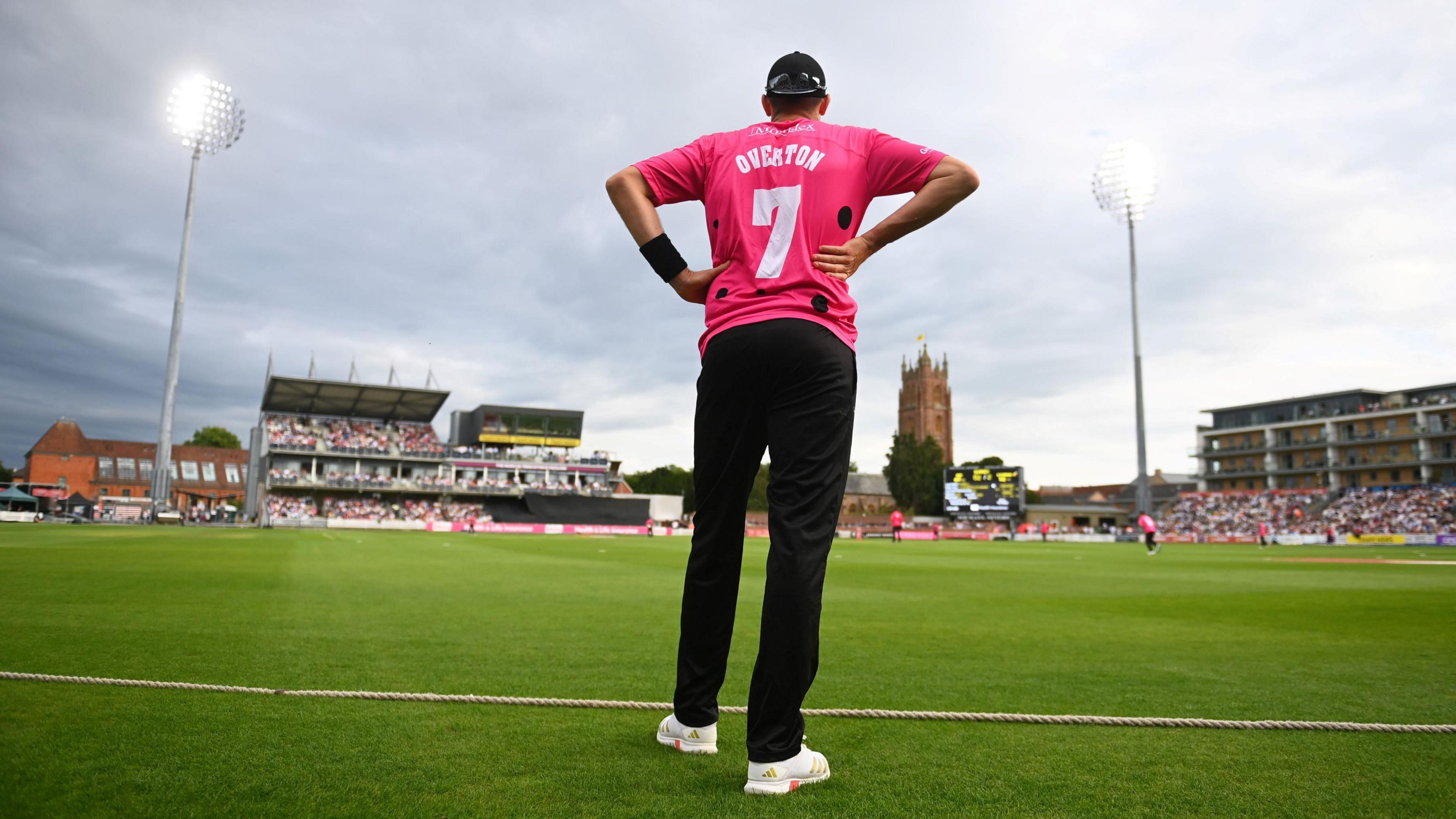 Somerset's Craig Overton watches from the boundary during the T20 match with Surrey at the county ground in Taunton. He is in the club's change kit of pink top and black trousers, and in the background packed stands and two of the stadium's floodlights can be seen