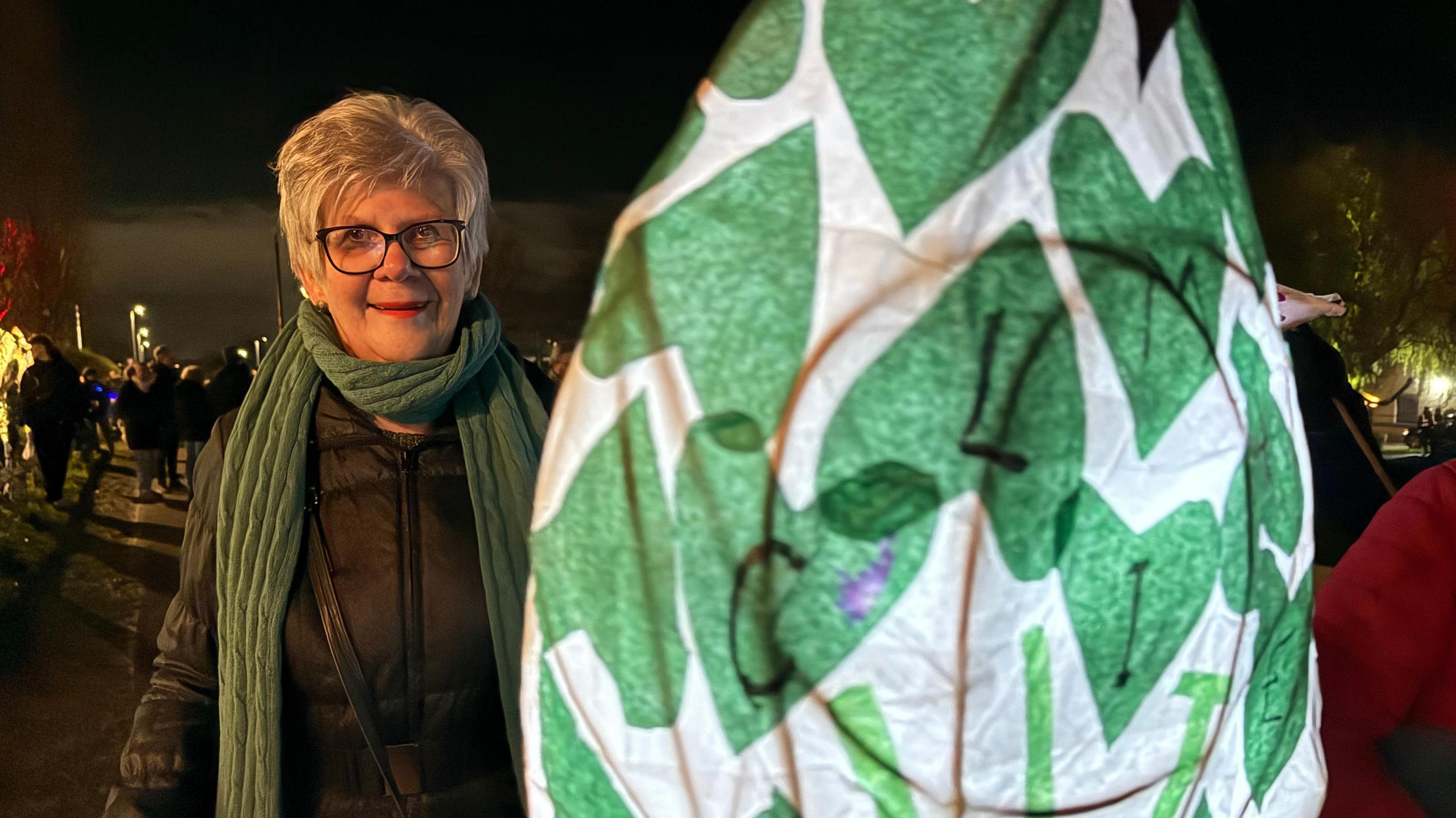 A woman wearing glasses and a green scarf holds up a green and white lantern.