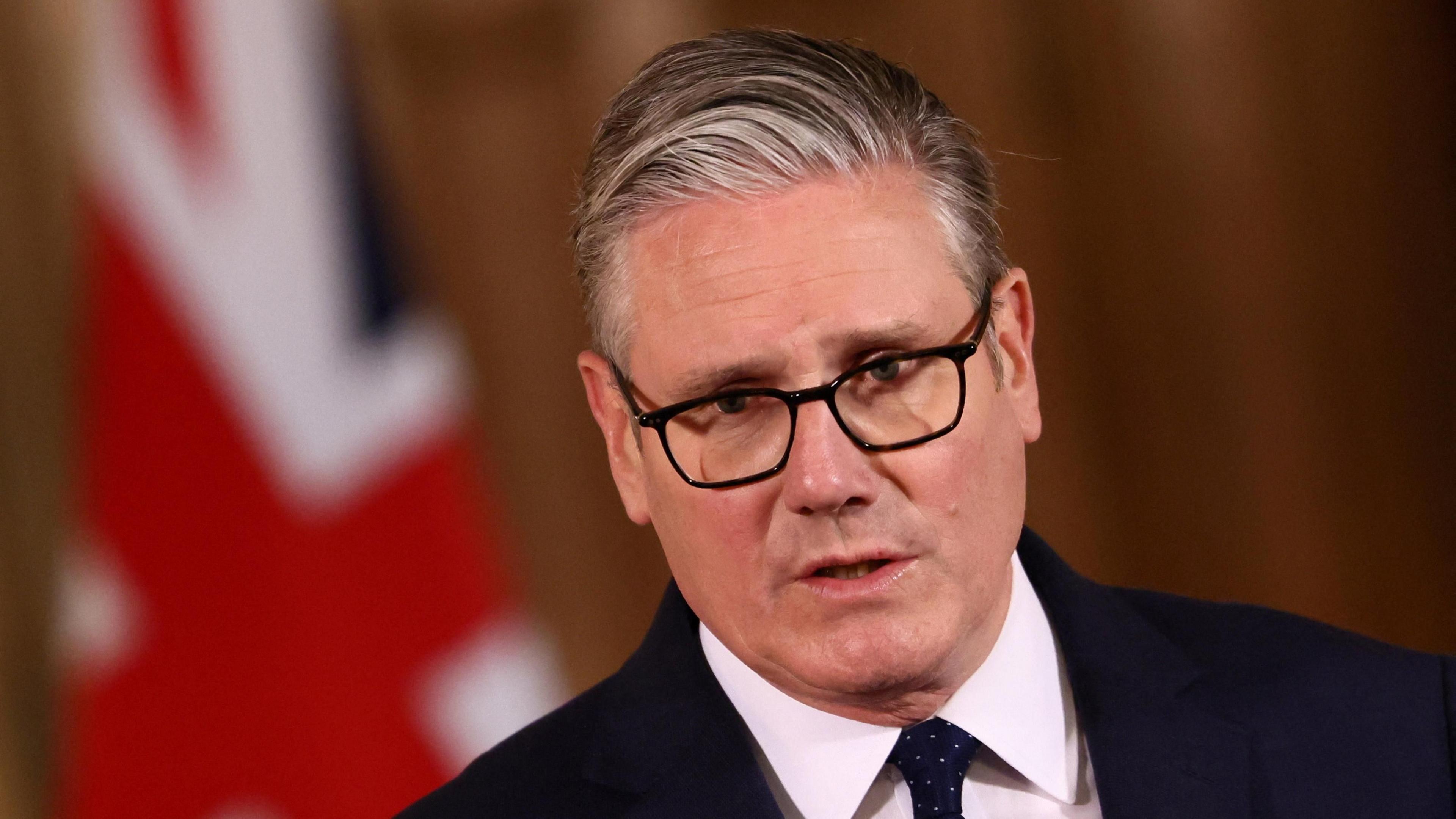 Sir Keir Starmer me kostum dhe kravatë të errët në një konferencë për shtyp në Downing Street, me sfond flamurin e Unionit
