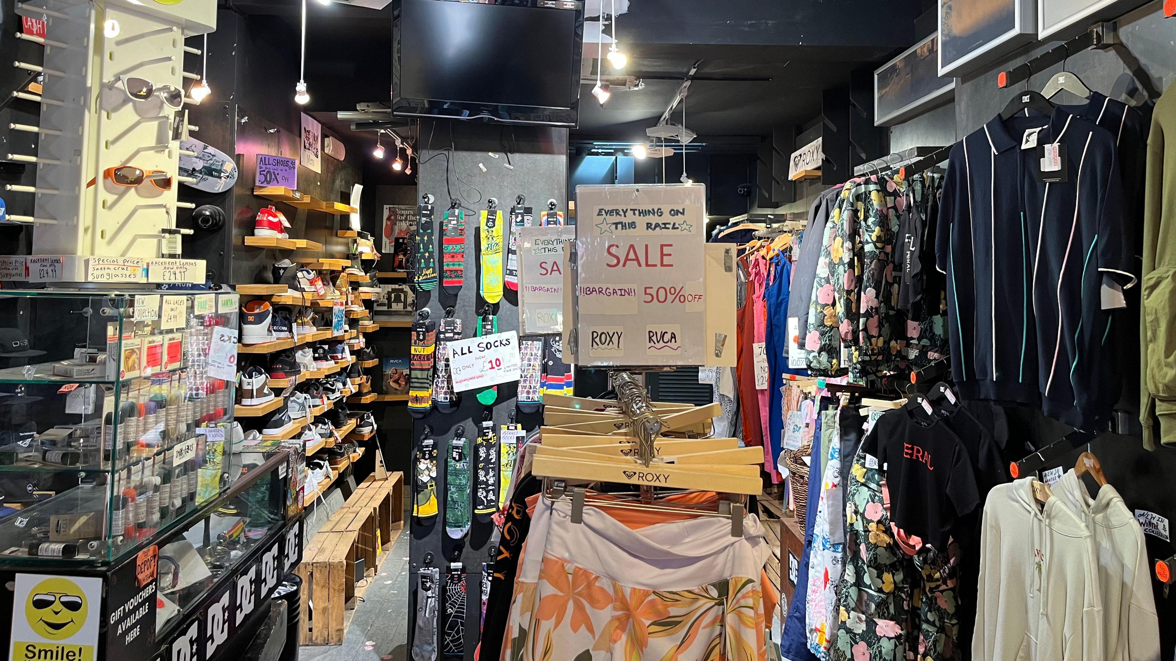 The inside of a skate wear shop. there are clothes hanging on the walls, as well as shoes and glasses. There are a number of signs reading 'sale' as the business is closing.