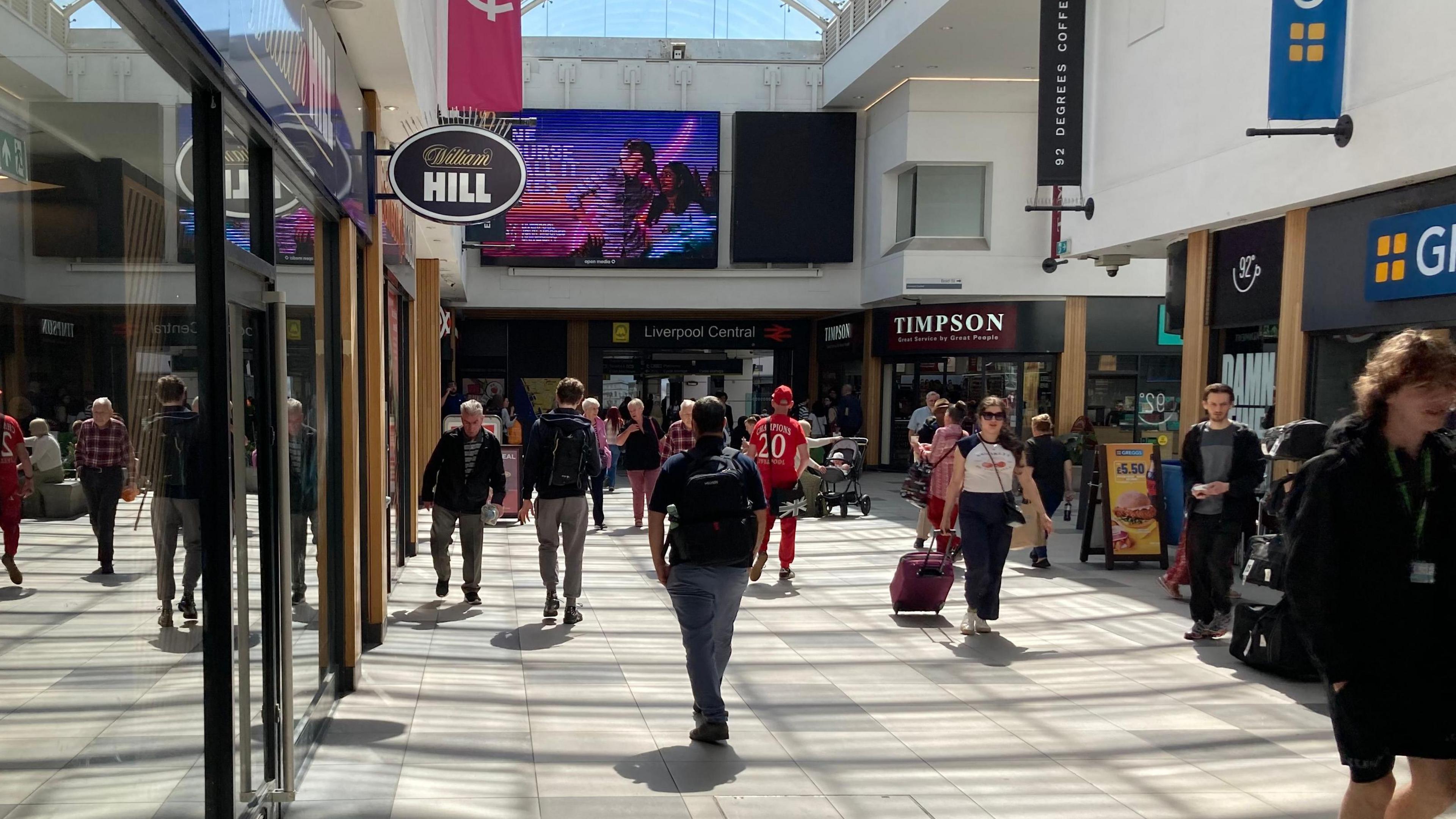 Inside Liverpool Central station concourse, with people shown walking towards and away from the station entrance. One woman is pulling a suitcase on wheels and a number of people are carrying rucksacks. The sun is shining through the glass roof and shadows are cast on the tiled floor.