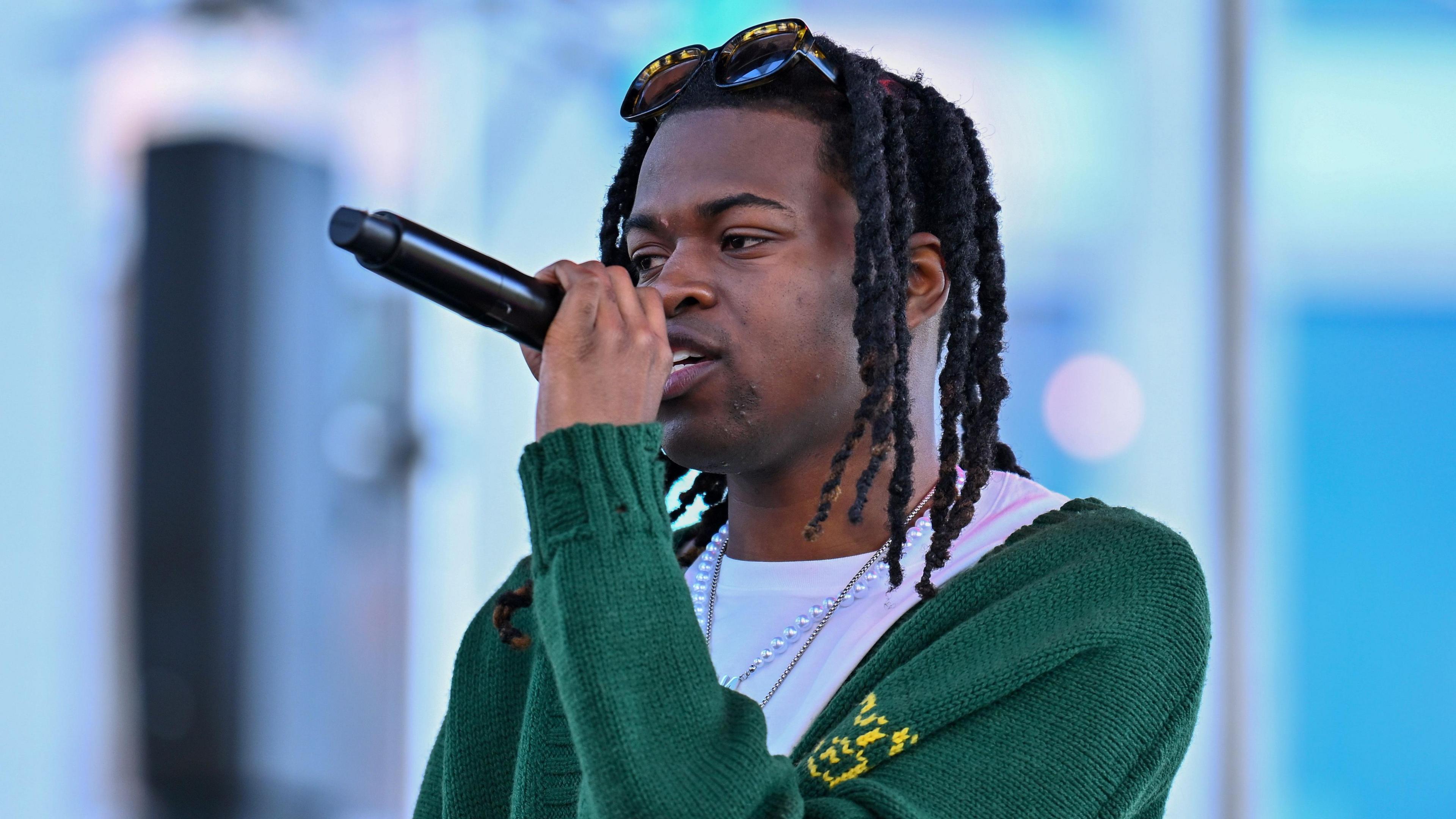 Jordan Adetunji is standing on stage performig. He's holding a mic and is wearing a green cardigan and a pearl necklace. There are sunglasses on top of his head. 