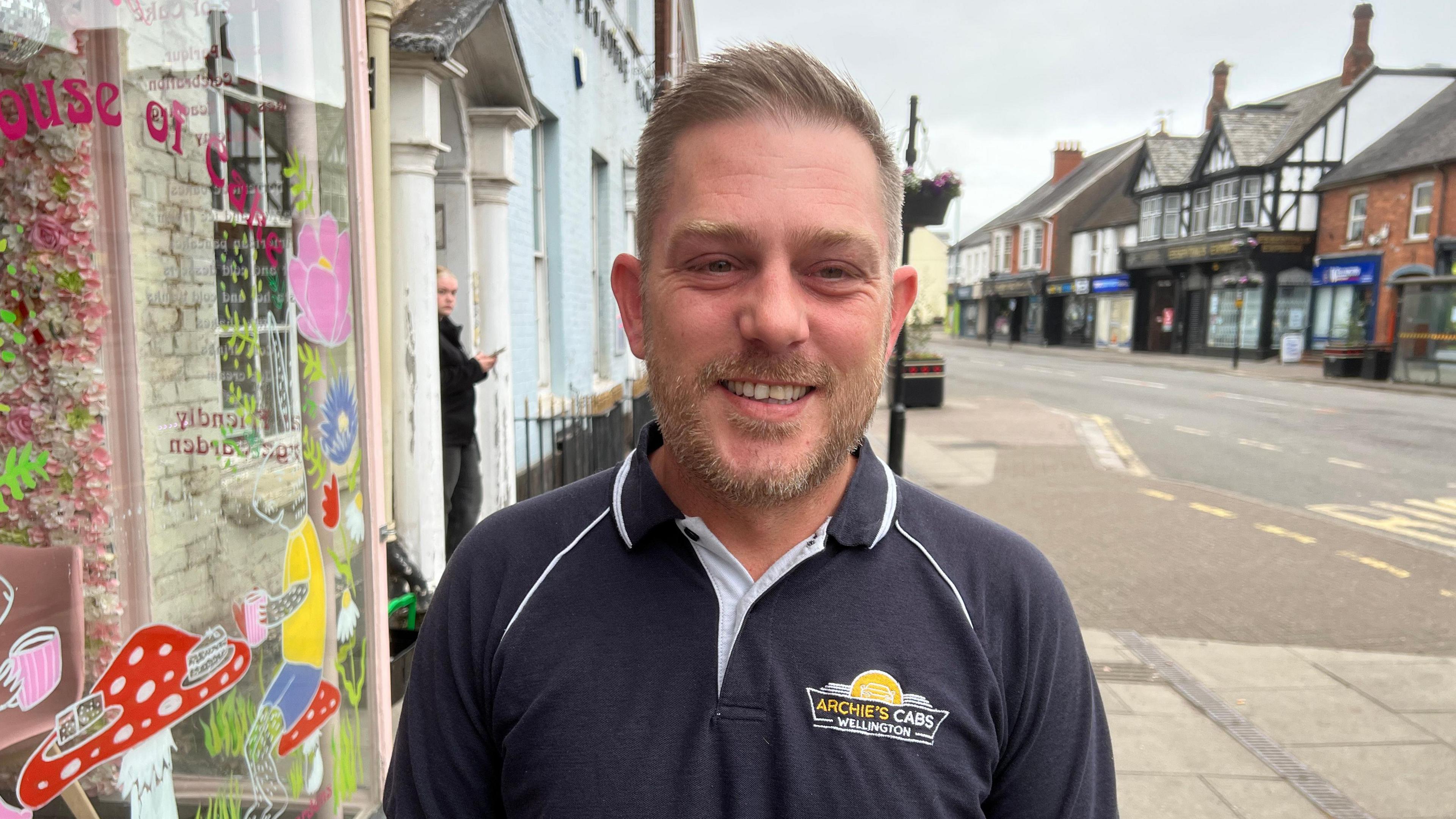 Paul Higginbottom is outside House of Cake in Wellington High Street. He is wearing an "Archie's Cabs" T-shirt.