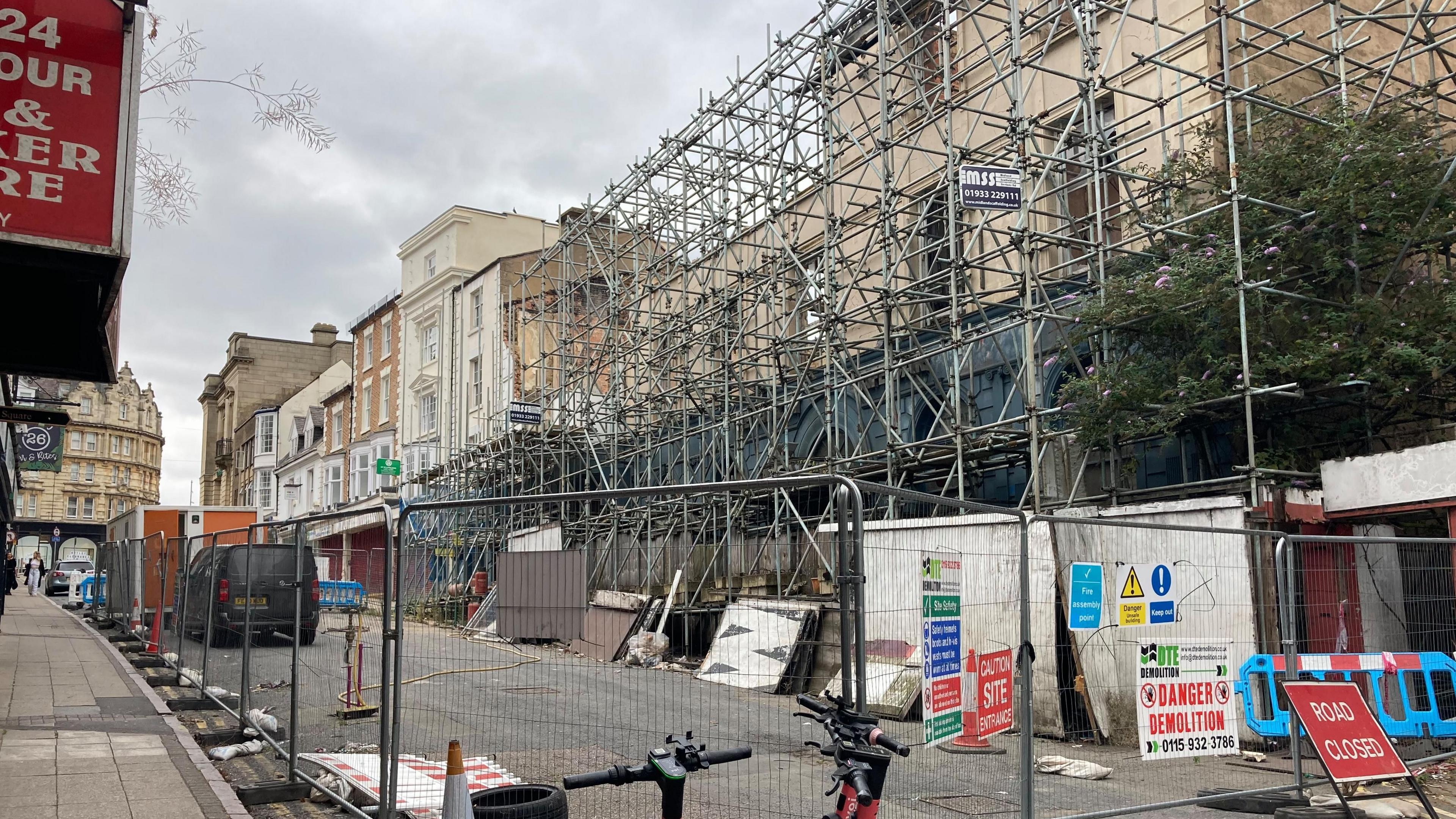 A street with silver barriers blocking the entrance to the road, with a red sign that reads "Road Closed". On the right is a large amount of erected scaffolding across a partly-demolished building.  