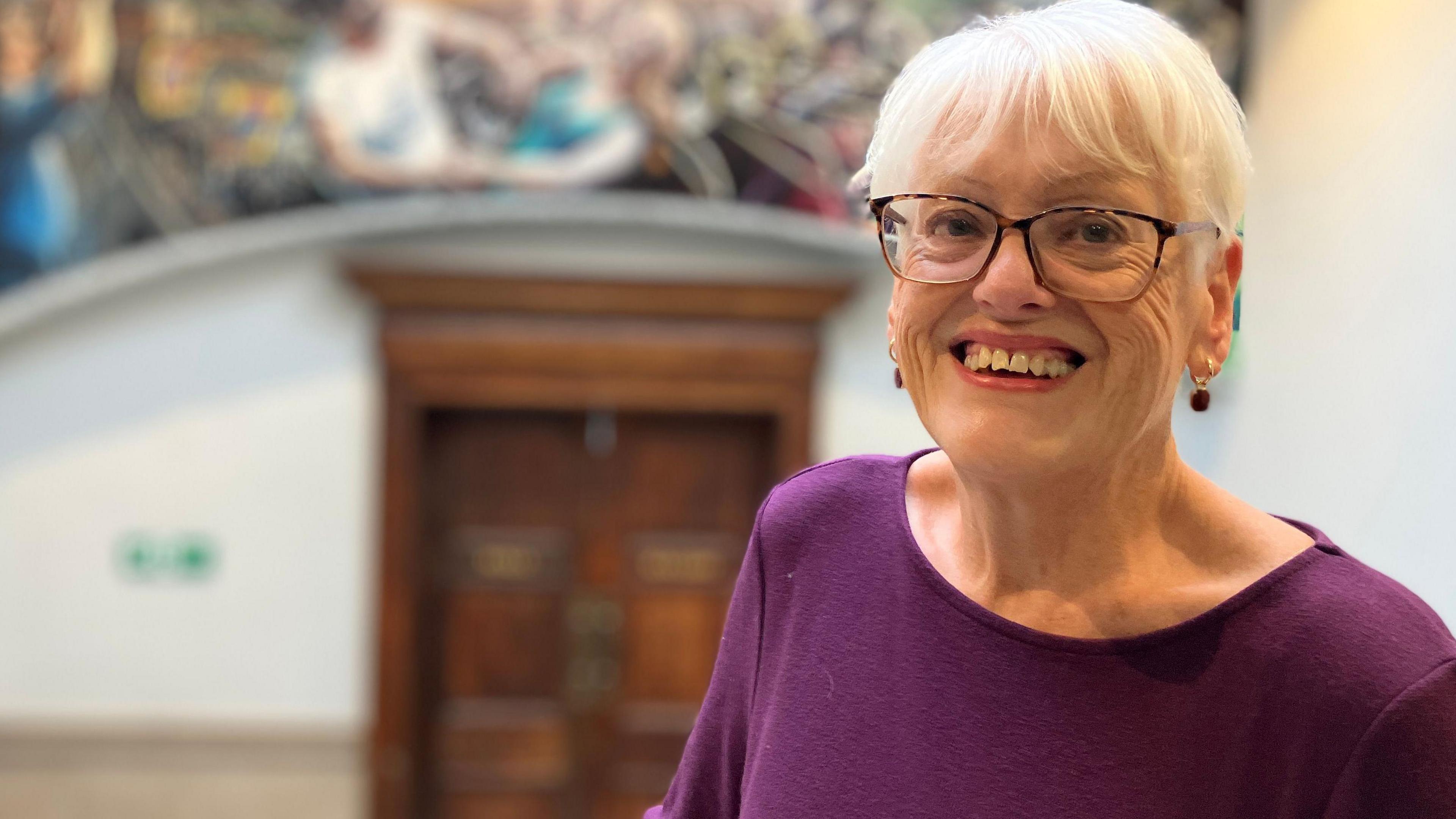 Mary Stratford smiling into the camera. She has white hair with a full fringe. She is wearing brown glasses and dangly earrings and a purple top. The background is out of focus, but she is standing in front of what looks like a large mural.