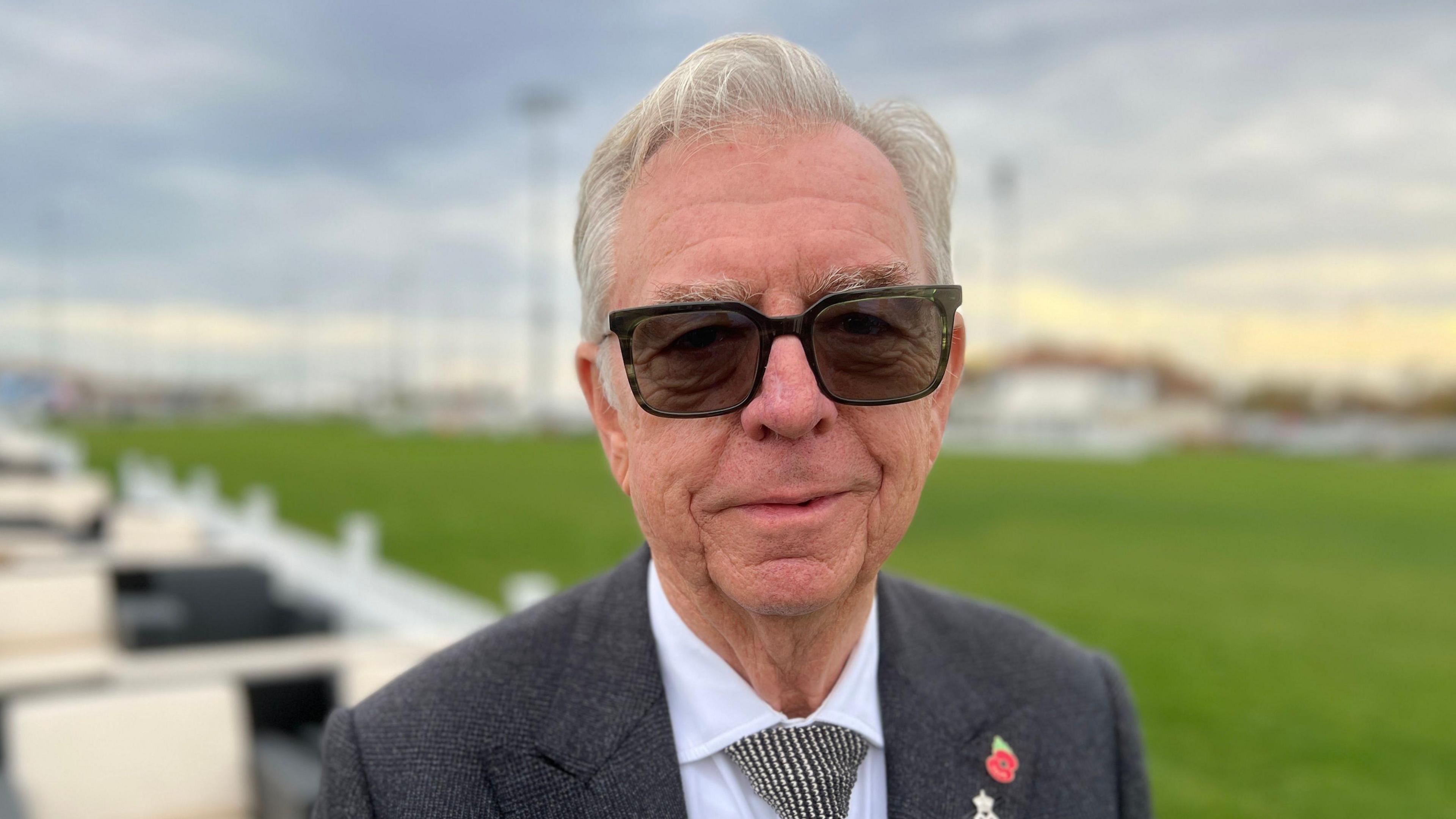 Philip Miller stands outside on a cloudy day in front of a large section of green grass. He has grey hair and wears sunglasses, a grey suit jacket, white shirt, black and white tie, and a small poppy pin on one side of his chest. 