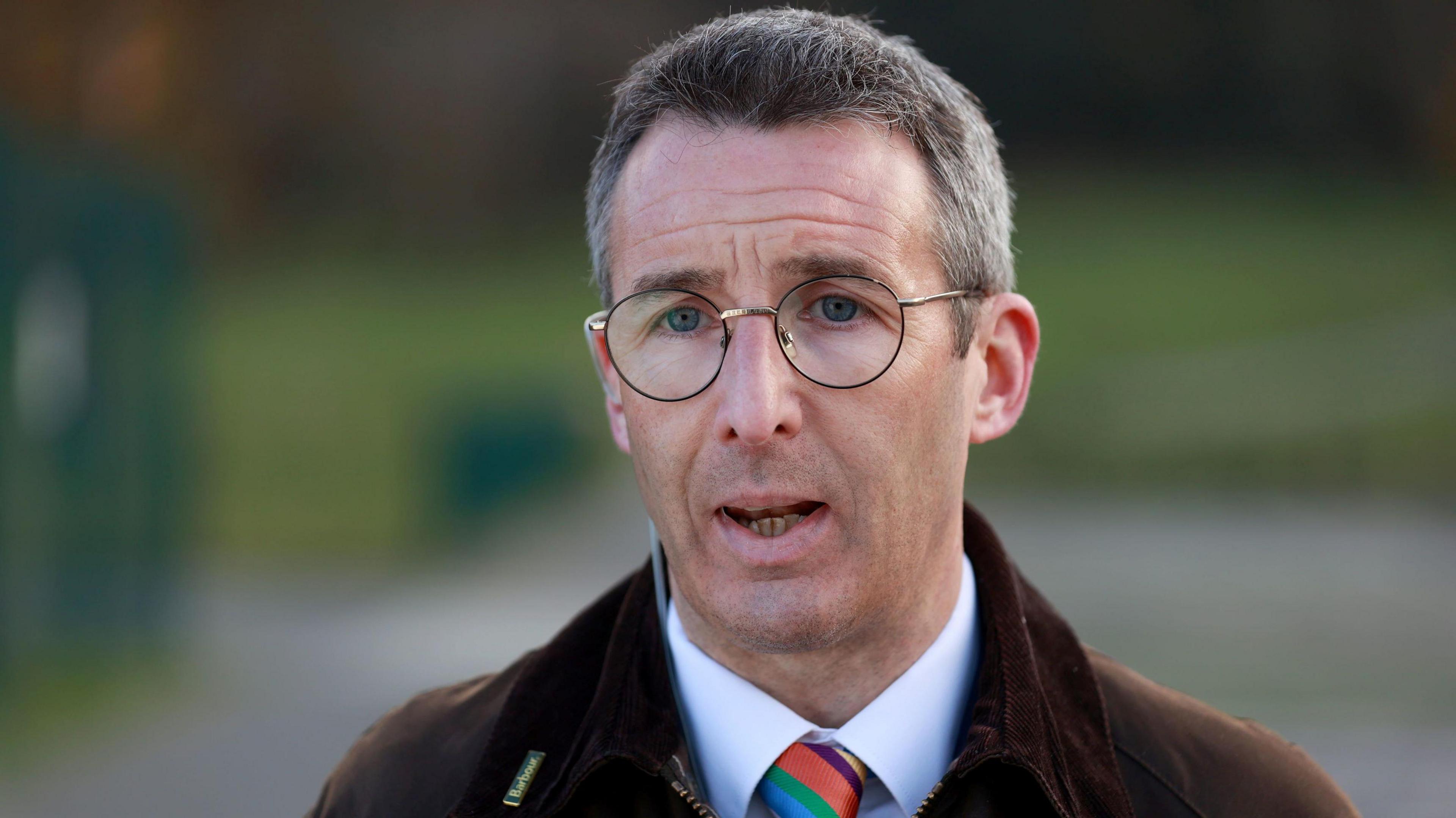 Andrew Muir looks at the camera, looking concerned. He has short, light hair and is wearing round glasses, a brown jacket, a white shirt and a colourful tie