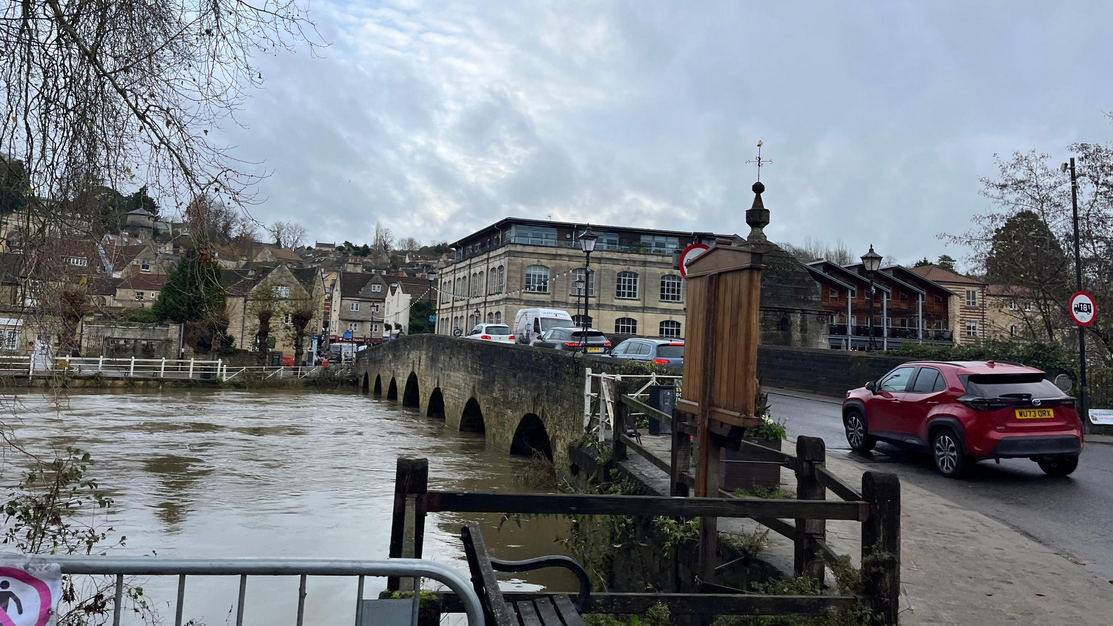 Cars at a standstill as they drive over the stone Town Bridge in Bradford-on-Avon, with brown water high up the bridge. 