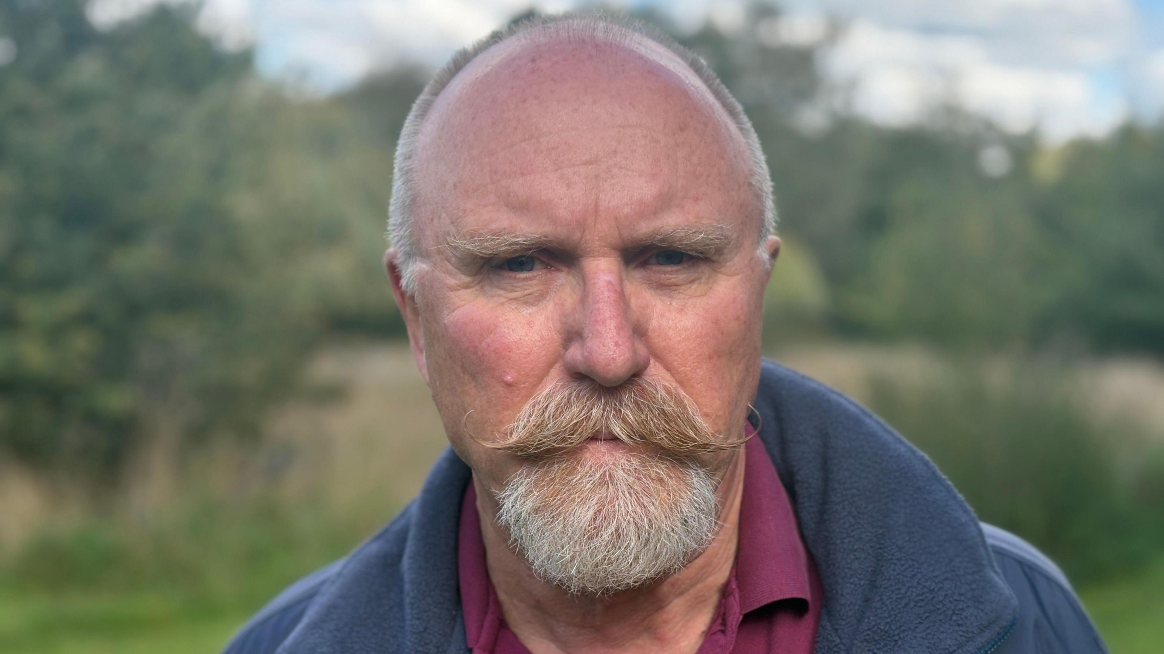 A man with short grey hair, a large moustache and a beard, and wearing a blue coat and burgundy shirt, stands in a field.