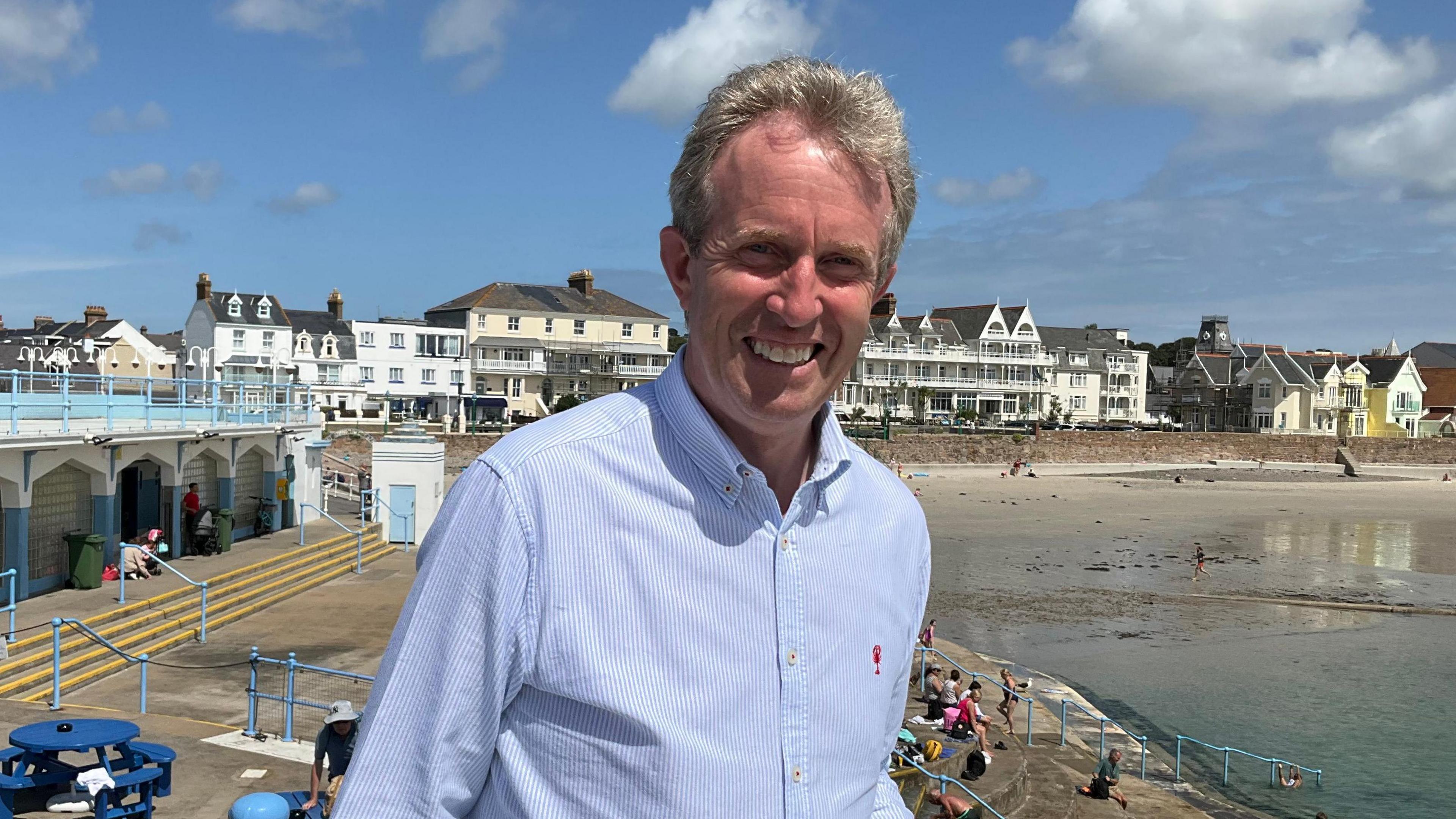 David Warr: A man with light grey hair smiling with teeth wearing a striped blue long sleeve shirt. Background of the lido, the beach and houses further afield, with light blue skies and a few clouds