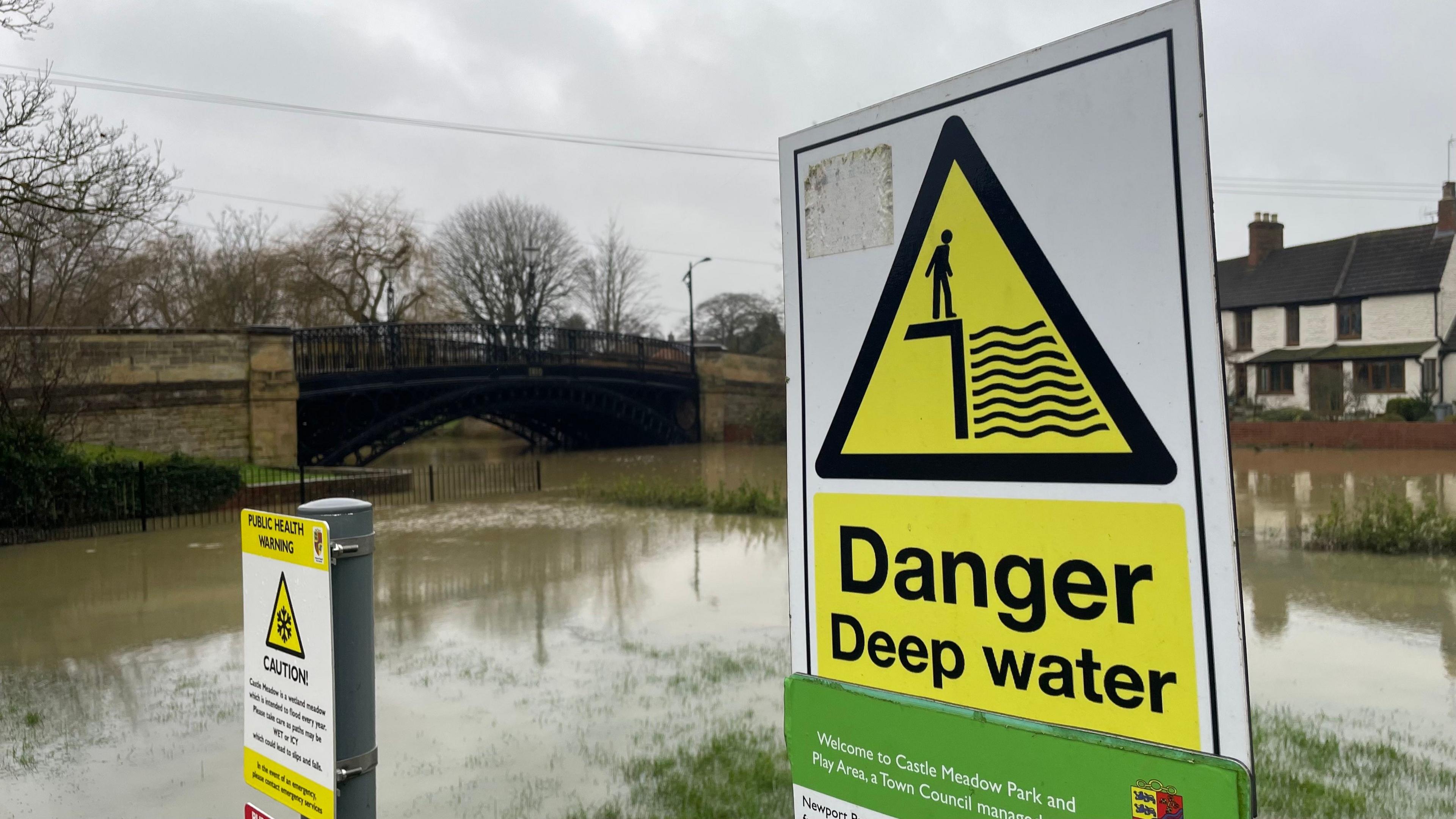 A sign saying 'Danger deep water' with a river and bridge in the background. A smaller sign on the left of the image explains the site is a water meadow that is designed to flood each year. The ground behind the signs is clearly flooded and is almost indistinguishable from the channel of the river.