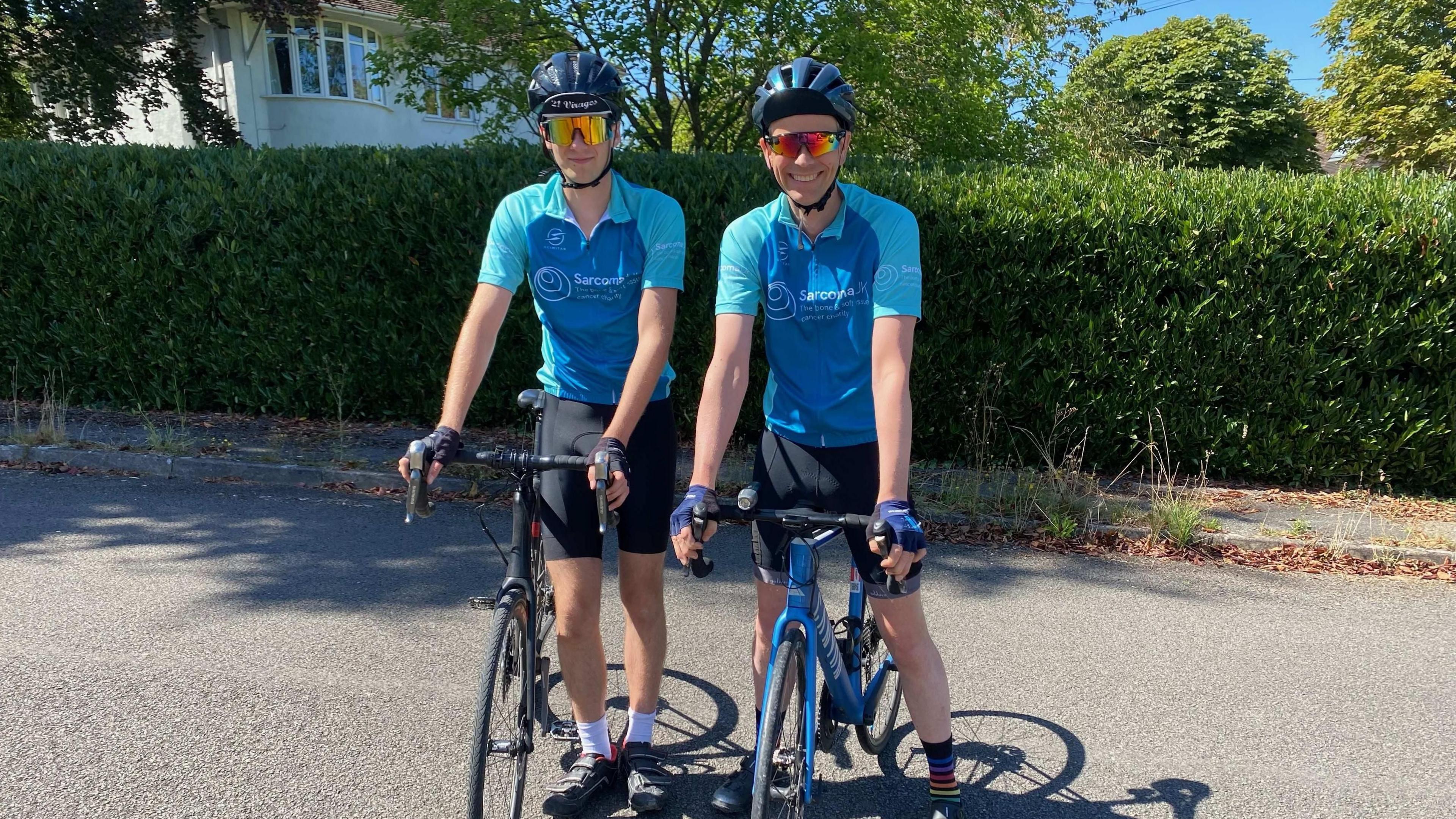 Jasper and Dominic are both straddling bikes, wearing cycling shorts and lycra turquoise tops with helmets and sunglasses.