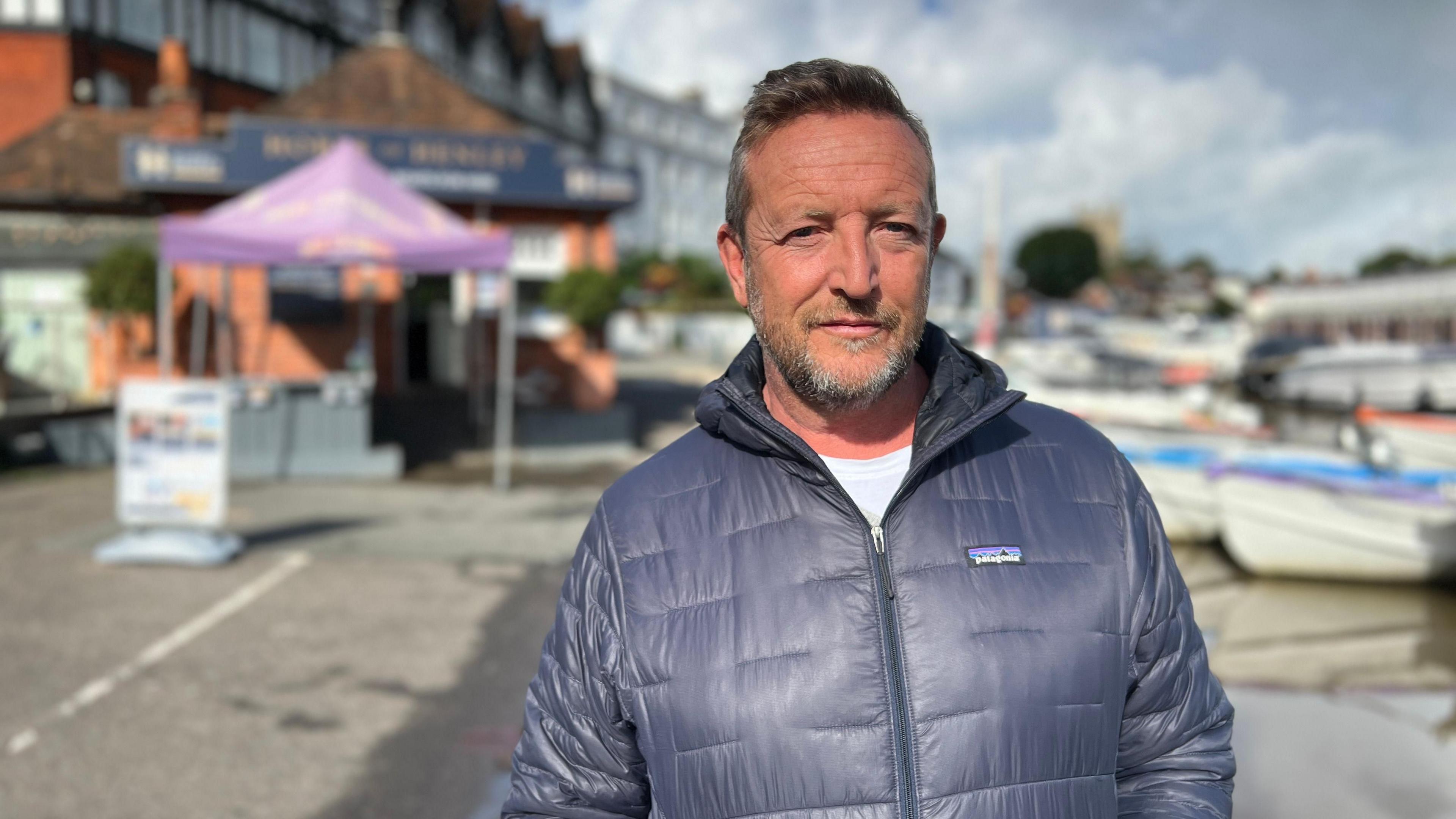 Jonathan Hobbs, with short blonde hair and stubble and wearing a blue waterproof coat, stands in front of a waterfront with boats lined up on one side.