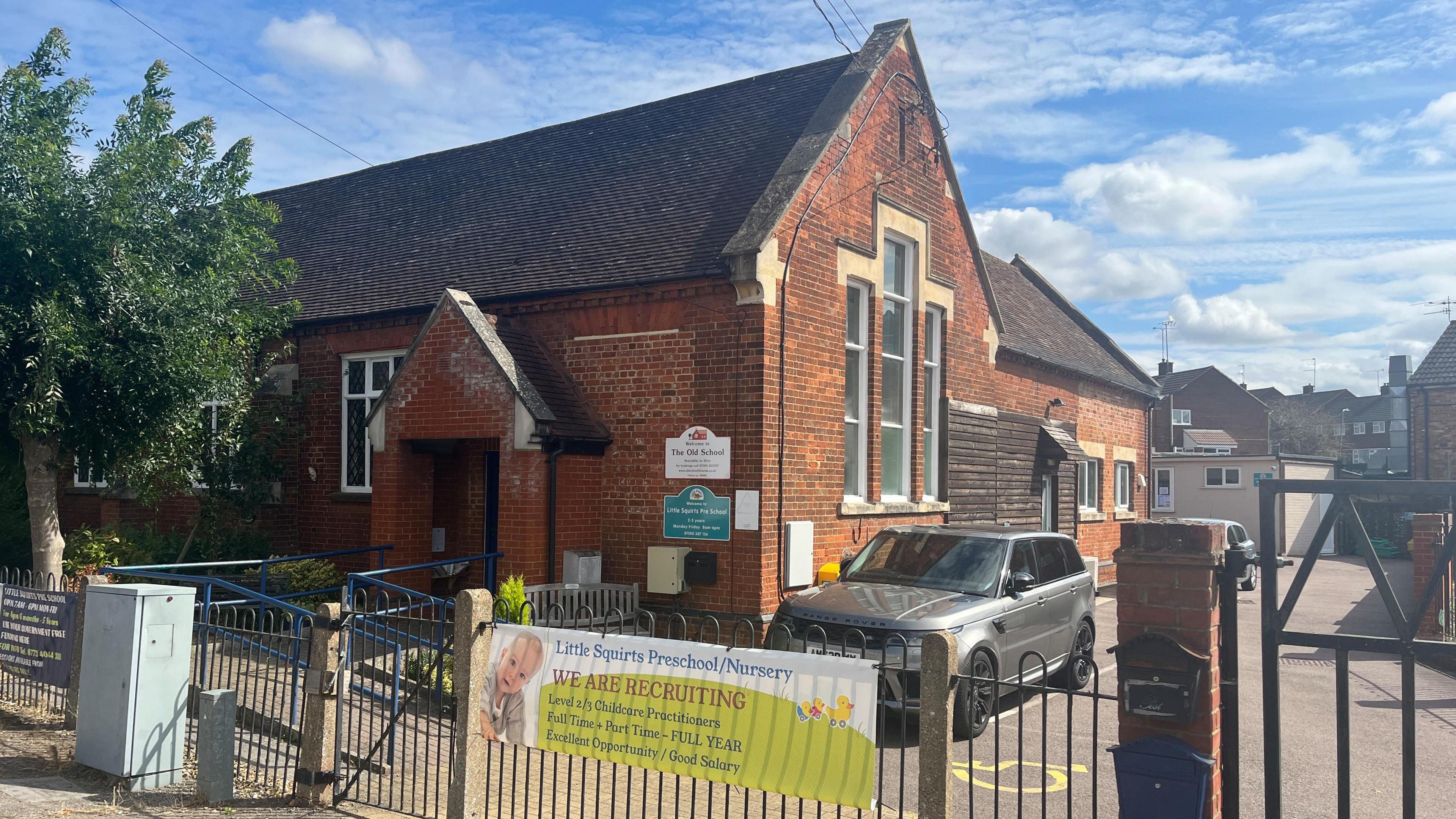 The old school building used by Little Squirts Preschool with a Range Rover parked outside on the other side of a metal fence