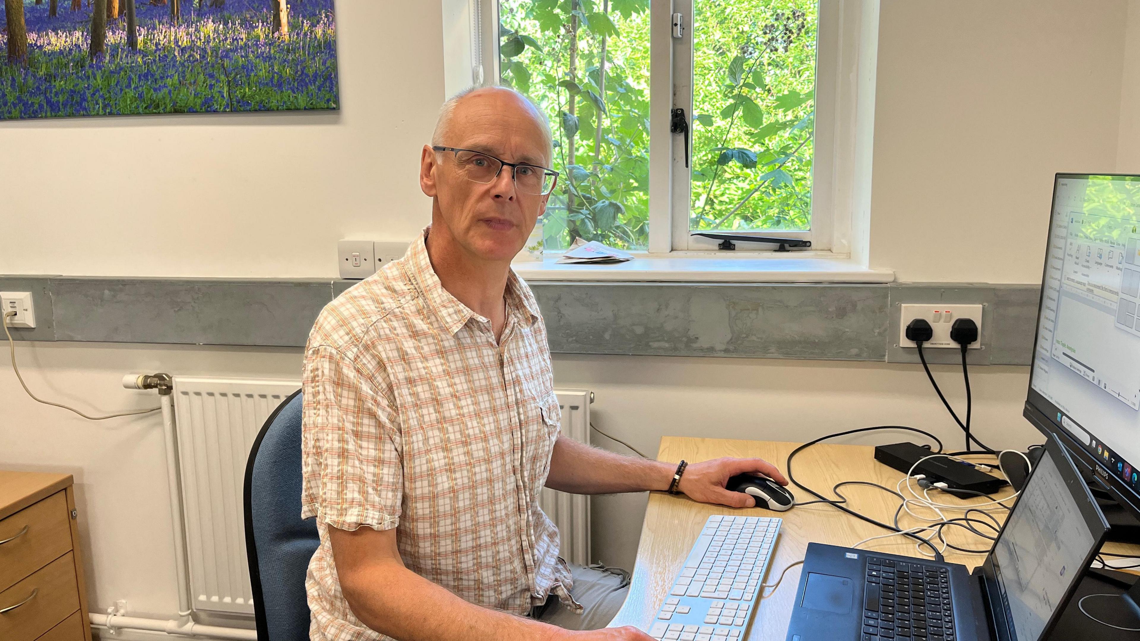A person sits at a desk in a modern office environment, working on a computer setup that includes a laptop and an additional monitor. Behind them, a window reveals a view of lush greenery, bringing a touch of nature into the workspace. A framed picture of a forest scene with blue flowers hangs on the adjacent wall. The desk is equipped with various electronic devices and cables.
