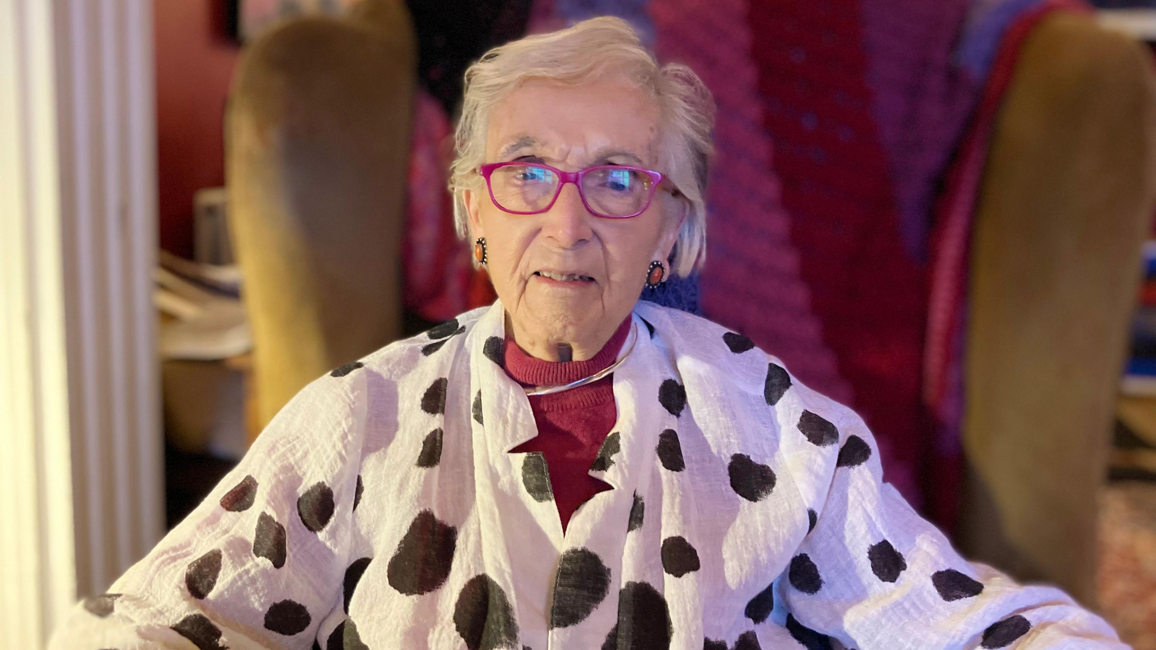 Glenys Cour sitting in a mustard armchair with a pink, red and purple blanket behind her. She has white short hair and is wearing pink glasses, pink earrings and a white blouse with black irregular spots. 