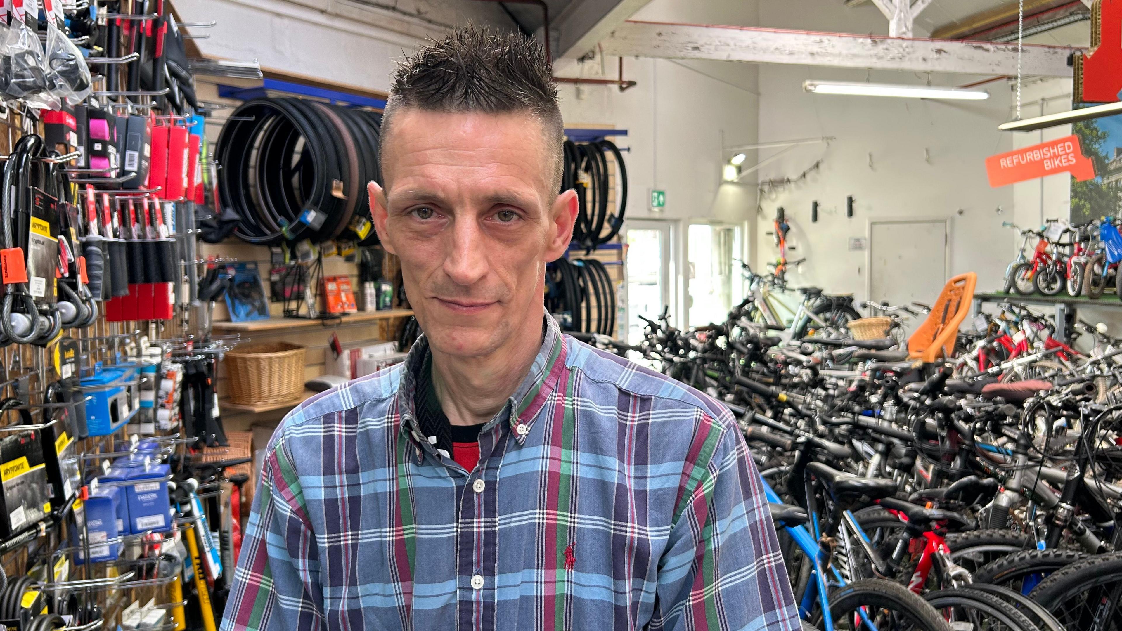 A head-and-shoulders picture of Simon Heard looking directly at the camera. He is wearing a blue checked shirt and has short hair which is shaved at the sides and gelled in small spikes on top. He is standing in front of lots of bikes in a workshop.