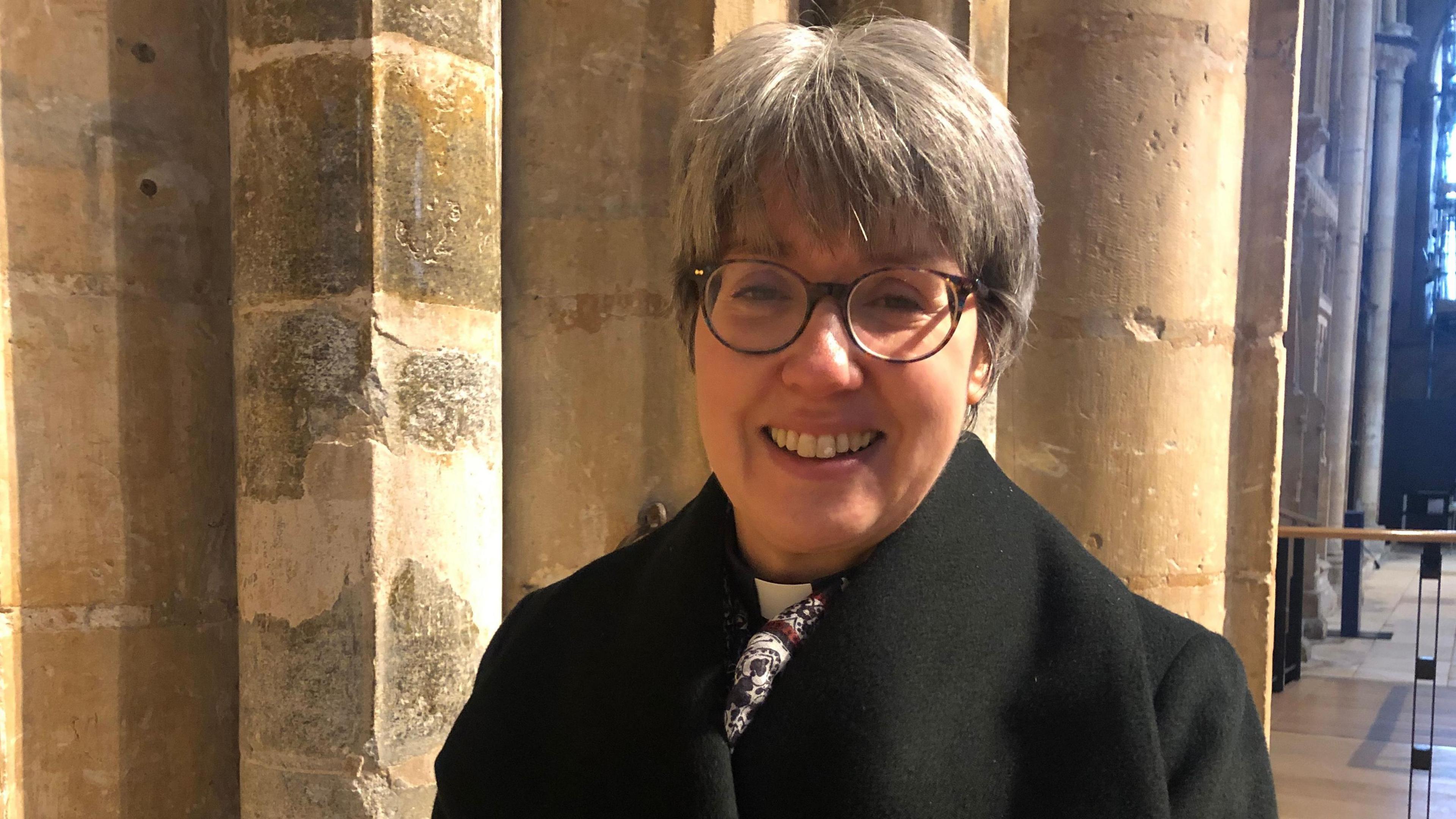 A woman, wearing glasses and a black coat, stands in front of stonework at Lincoln Cathedral.