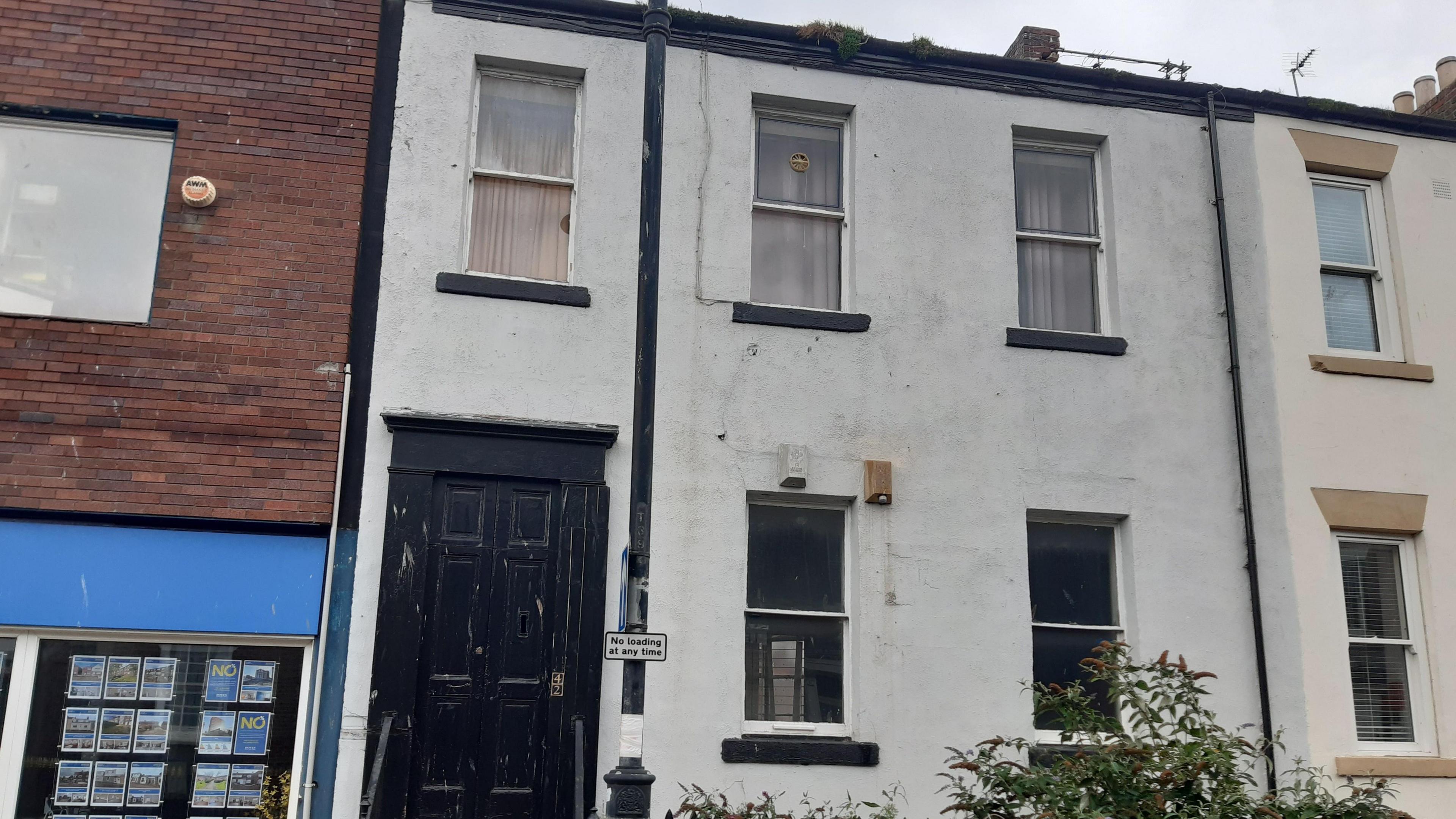 Terraced two-storey house with black door and grey paint on its walls. It has three windows on the top floor and two on the ground floor.