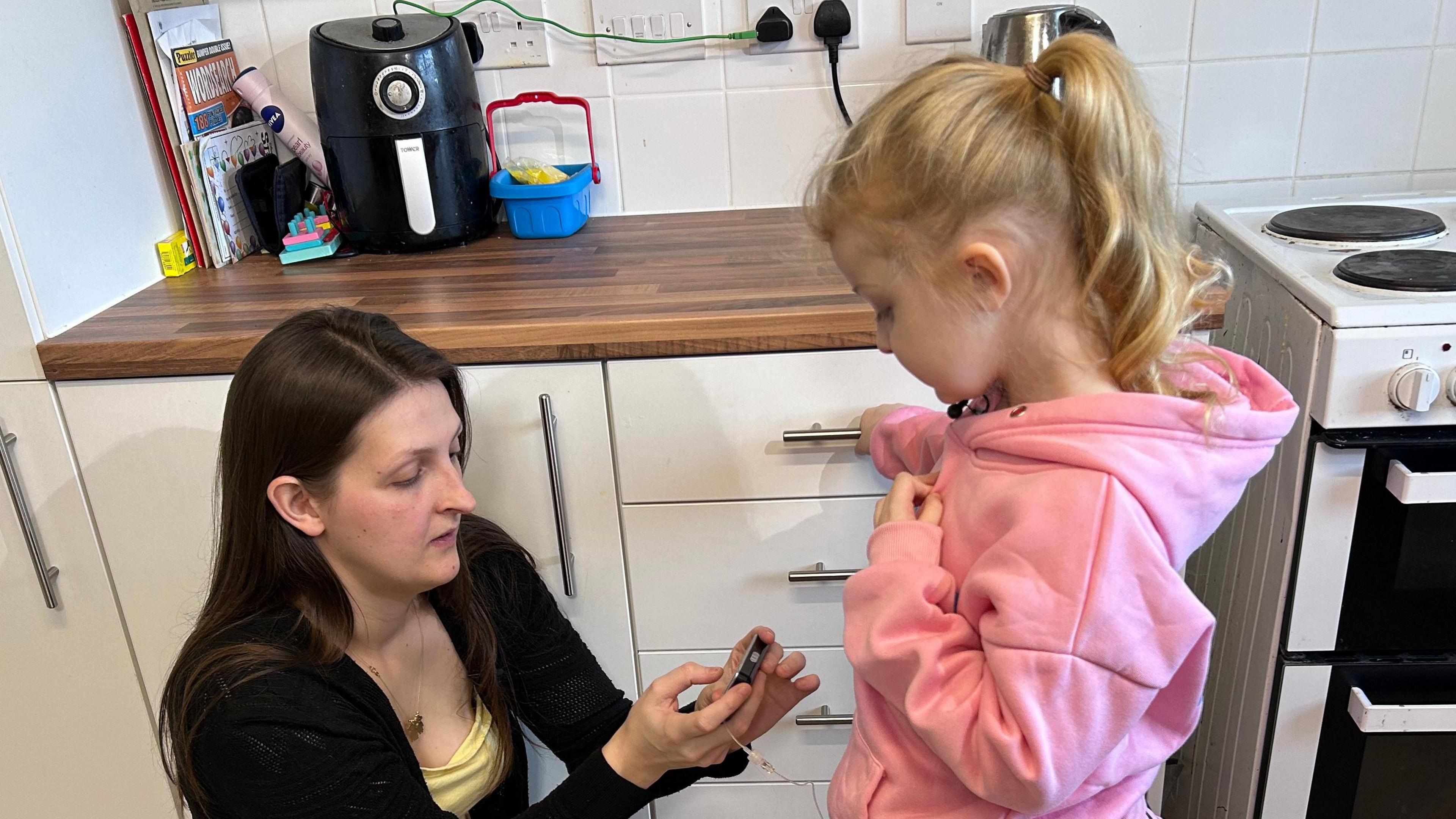 Lauren and Maisie look at her pump. Maisie is in a pink sweatshirt and Lauren is crouched down looking at the pump. She is in a black cardigan and yellow top. 