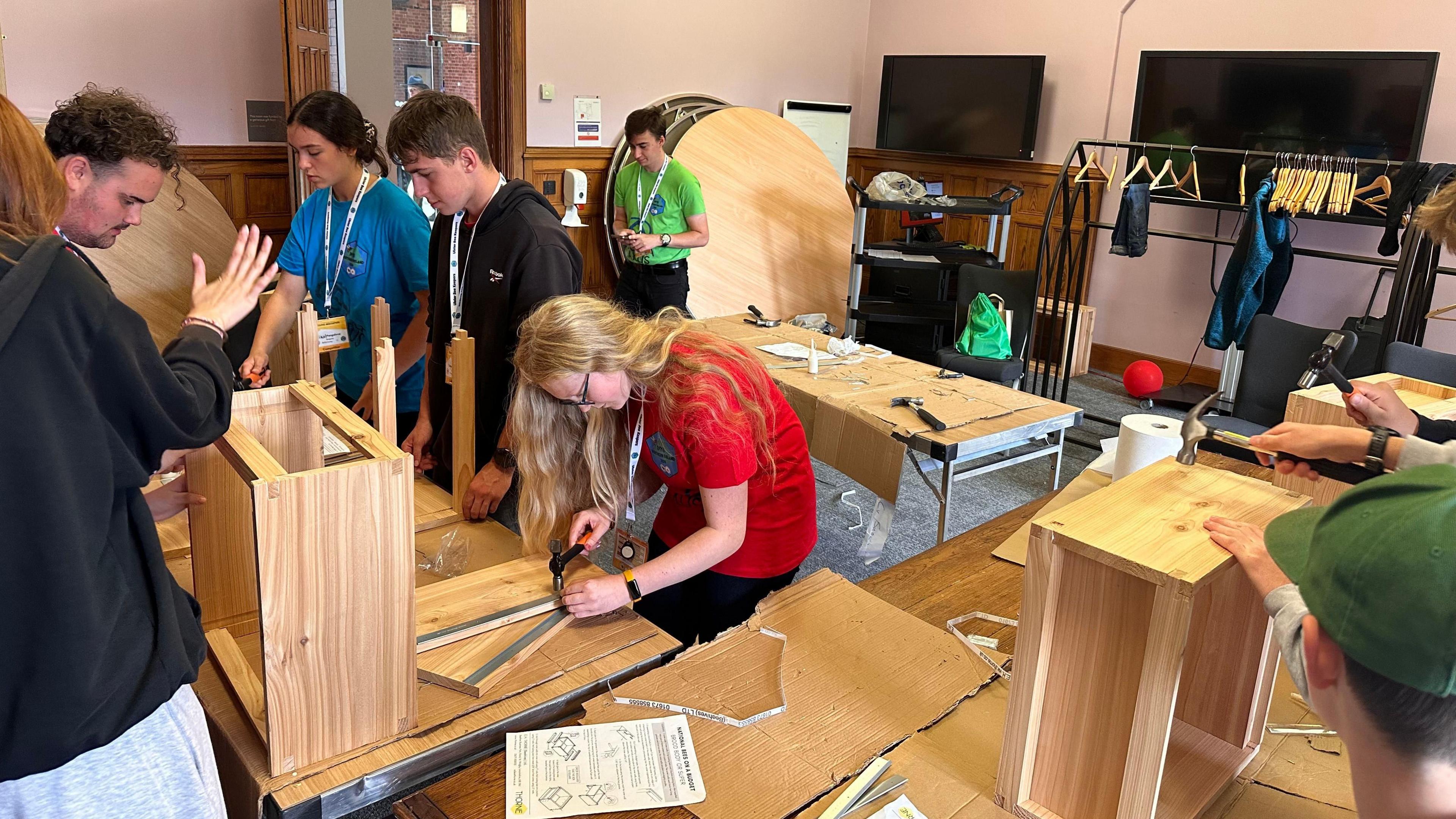 Young people using hammers and glue to make super boxes for bee hives. There are seven of them in the room.