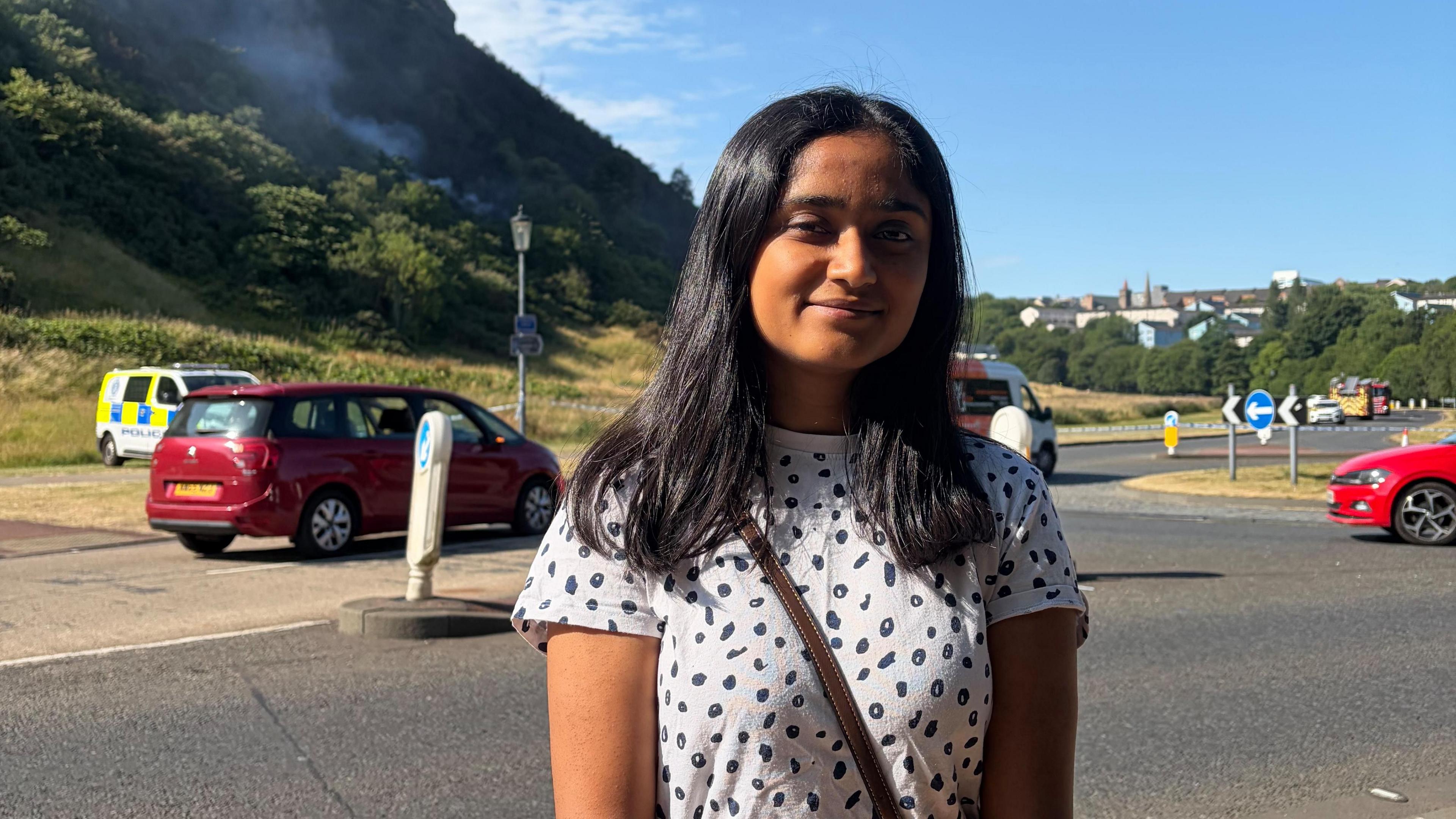 Mahek Jayantilial stands in front Arthur's Seat. She has dark hair and is wearing a black and white spotted t-shirt. 