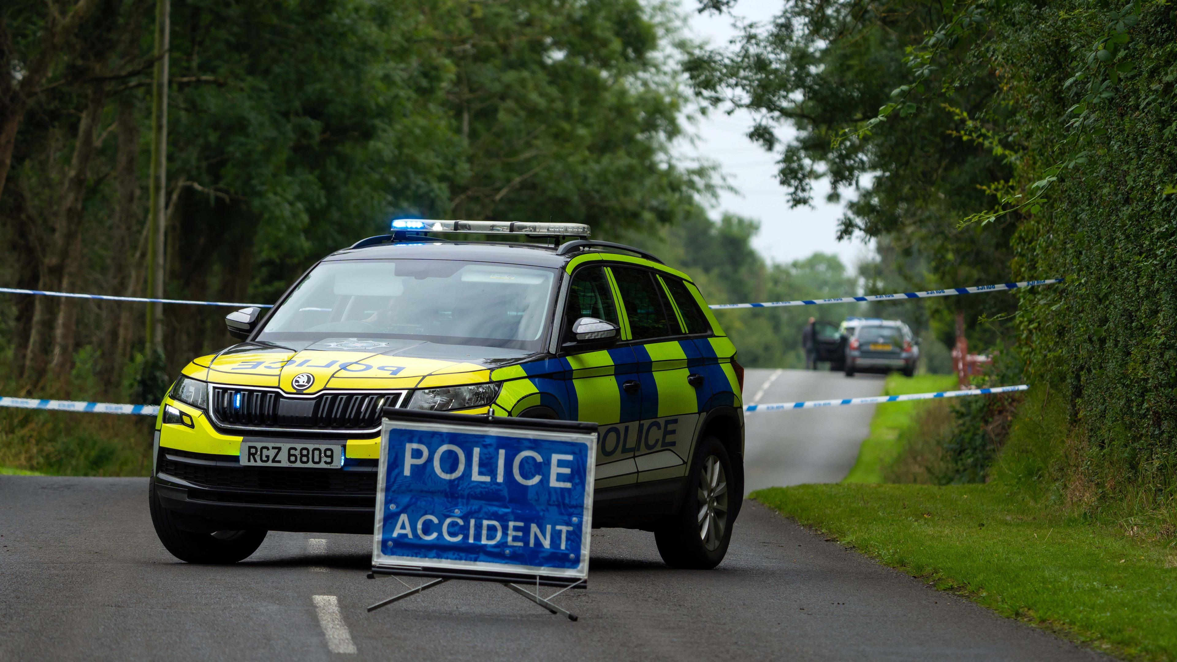 A blue sign in the middle of a road which reads POLICE ACCIDENT. There are trees and grass verge on side of the road. Behind the sign is a police car which has blue lights with a tape behind.