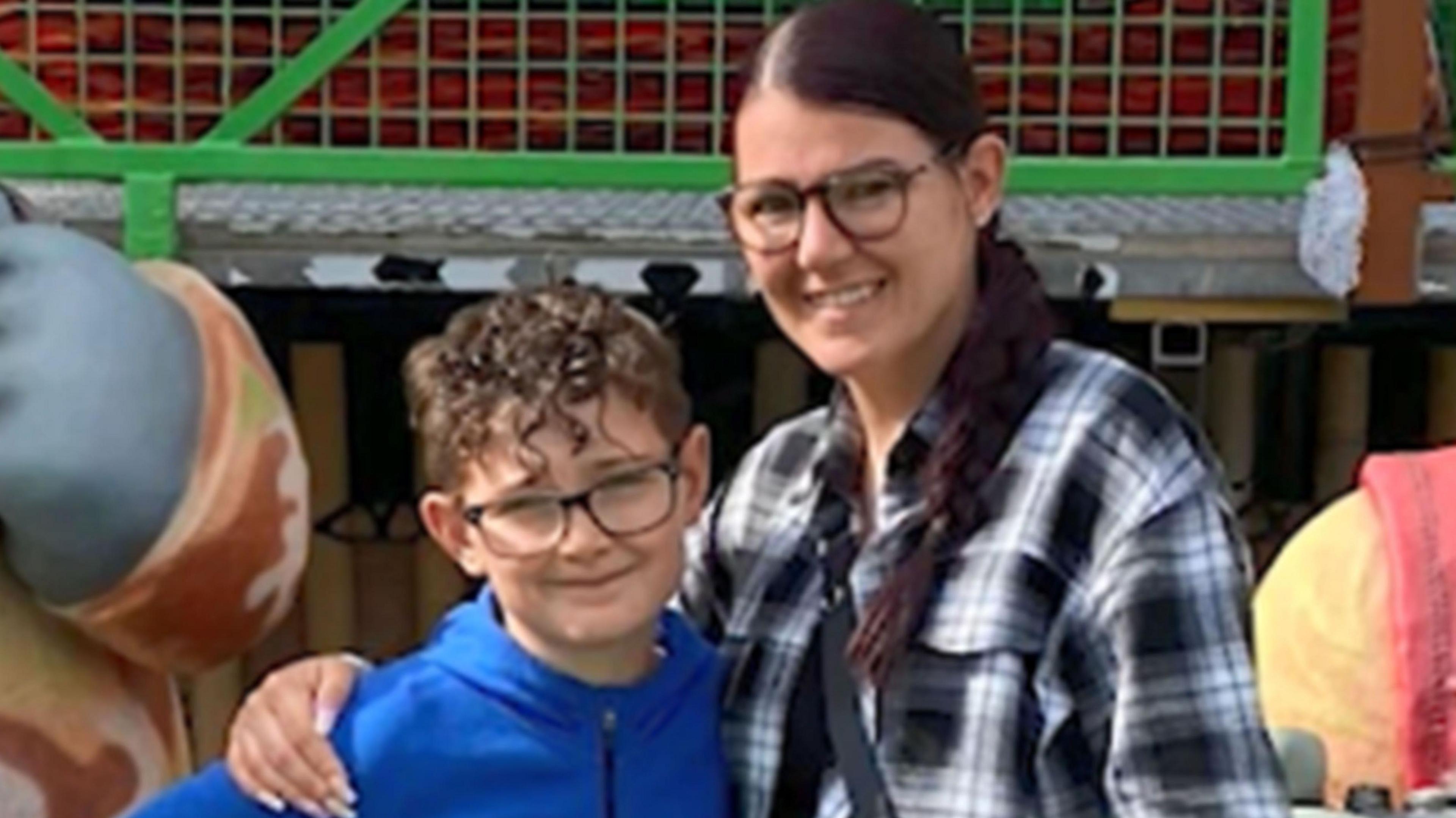 Jacob Calland, who is wearing a blue top and glasses, is pictured with his mother Carly Calland. They are standing in a theme park. 