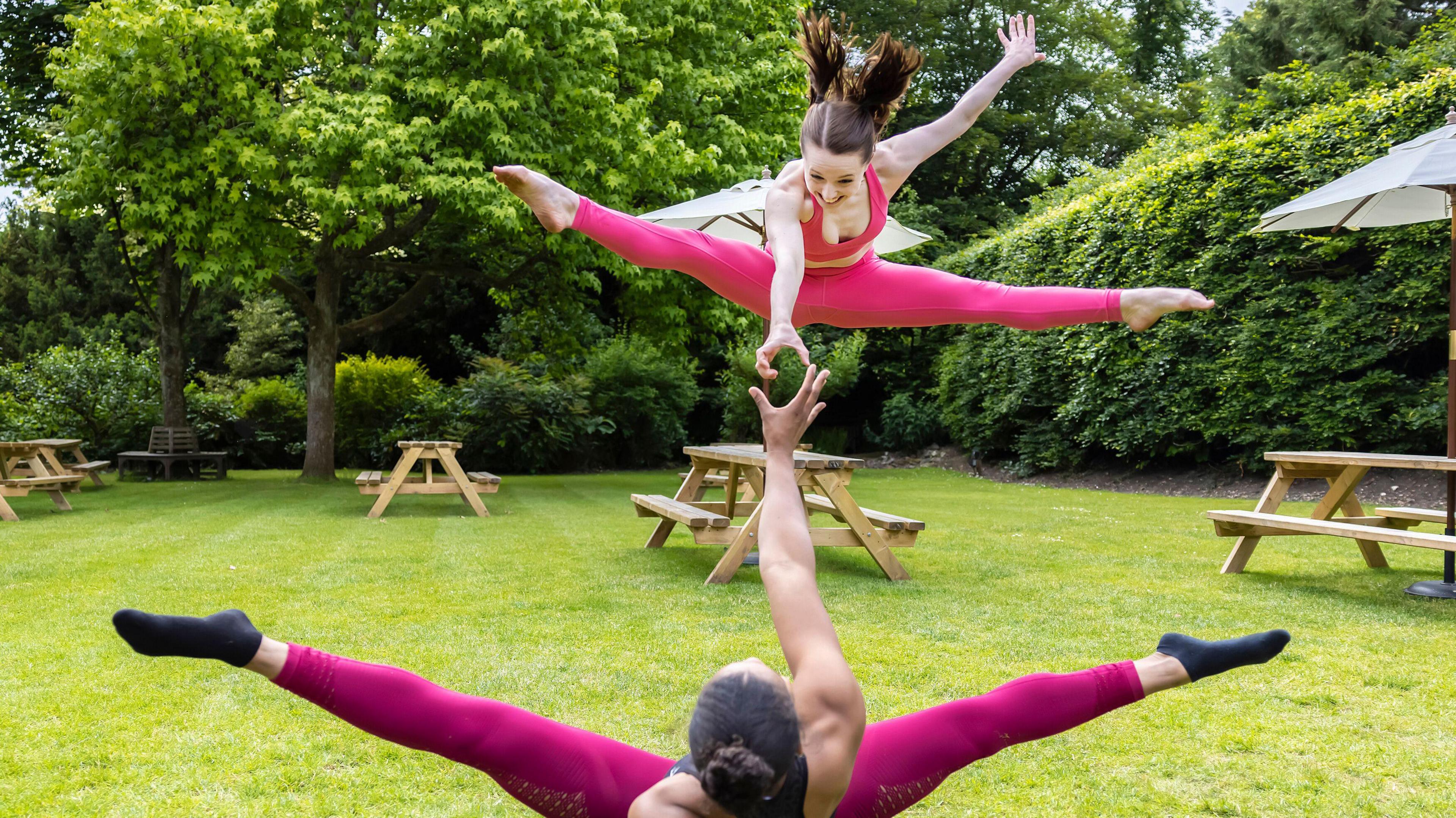 Two acrobatic performers, both wearing pink, one up in the air legs outstretched being caught by another on the floor