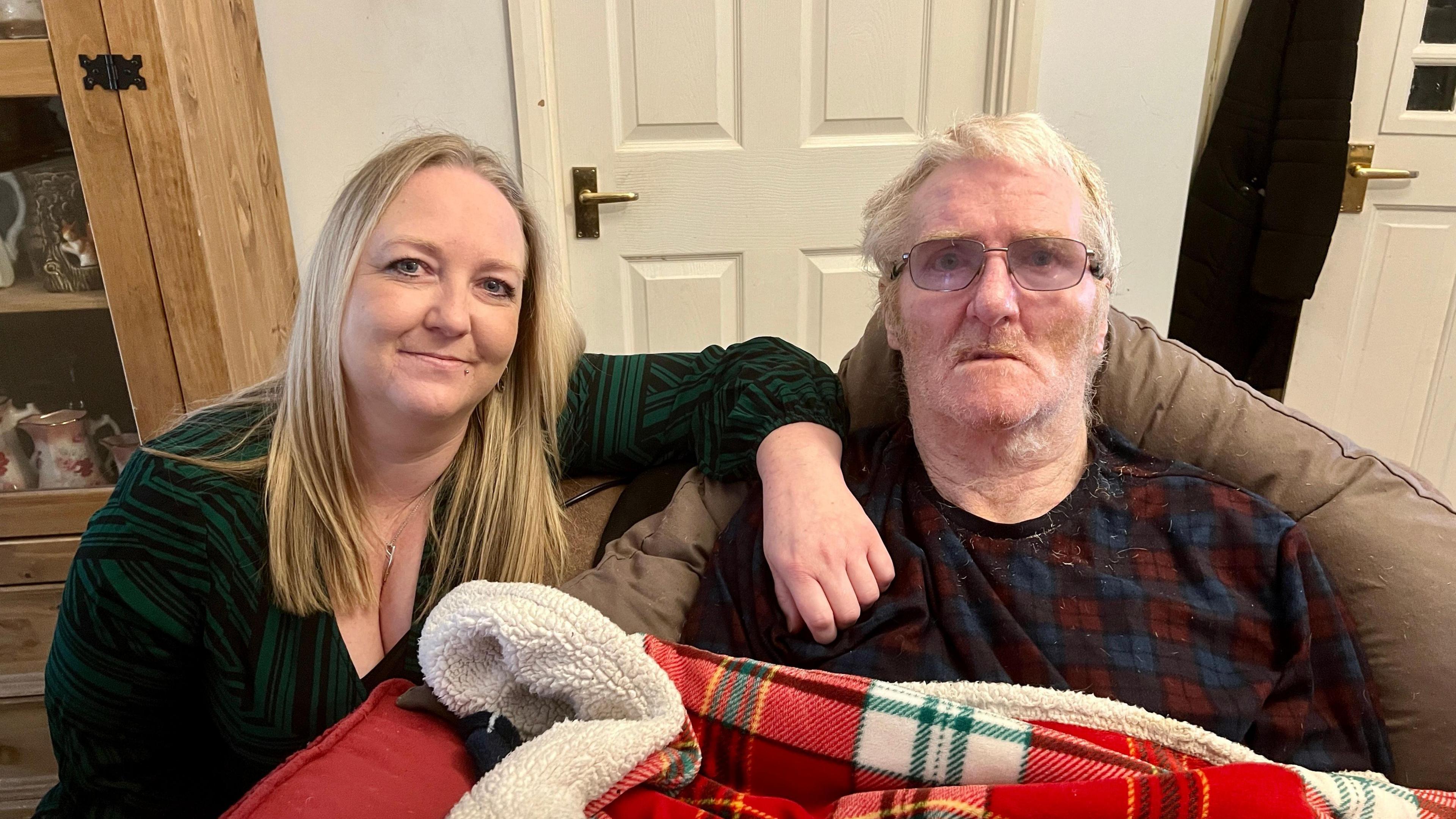 Rebecca crouches beside her father Rob who is sitting in a chair in the living room with a blanket over him. Rob struggles to talk because of his condition. He is wearing glasses and a checked T-shirt, he has grey hair but is in his early 70's. His daughter has long blonde, straight hair and is wearing a black scoop neck top.